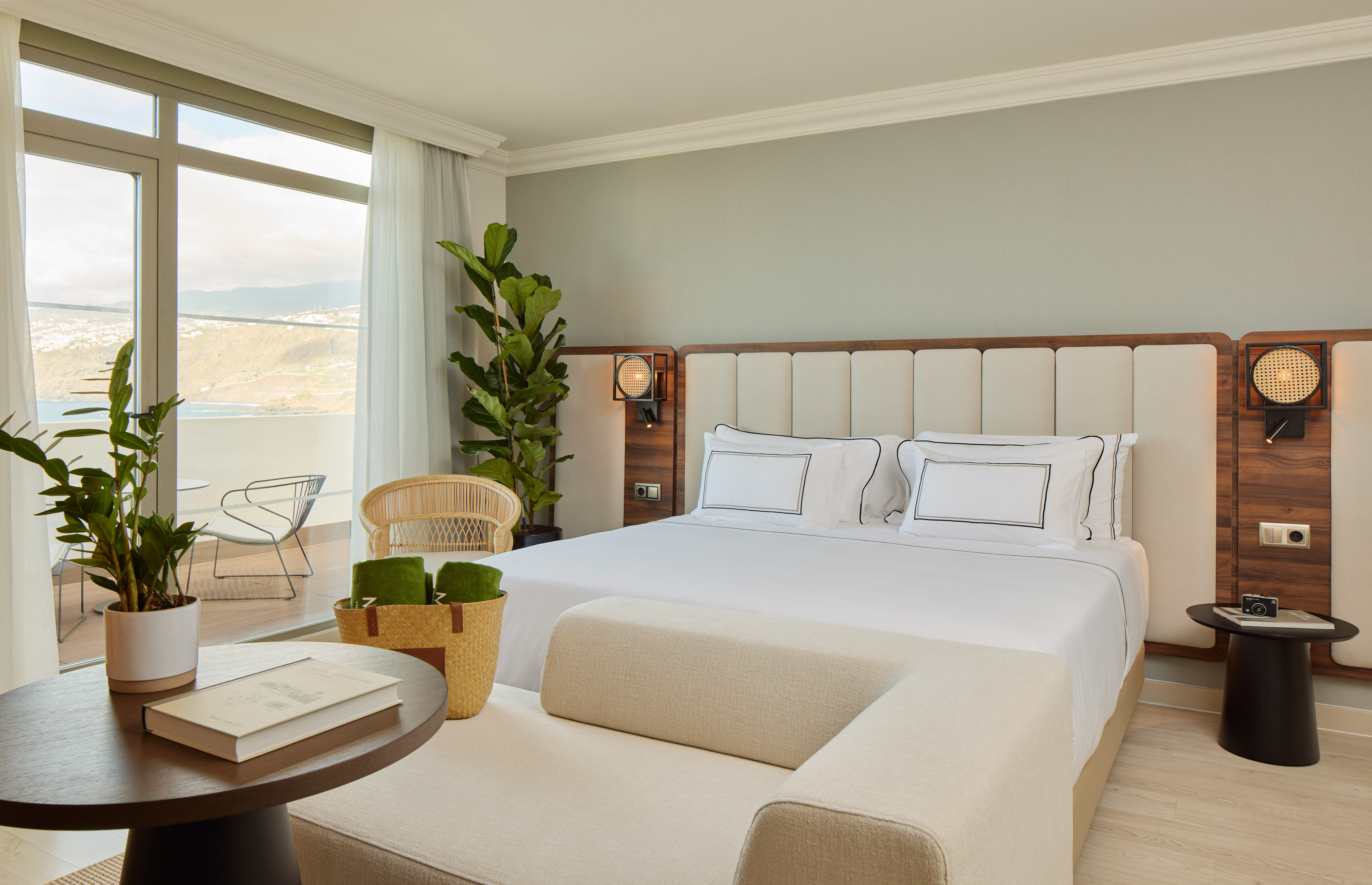 Modern hotel bedroom with white bed, balcony ocean view, and green plants.