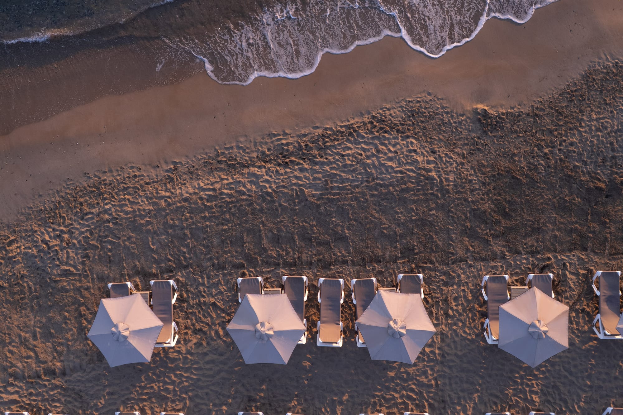 Aerial beach view: rows of grey umbrellas, sun loungers, ocean waves.