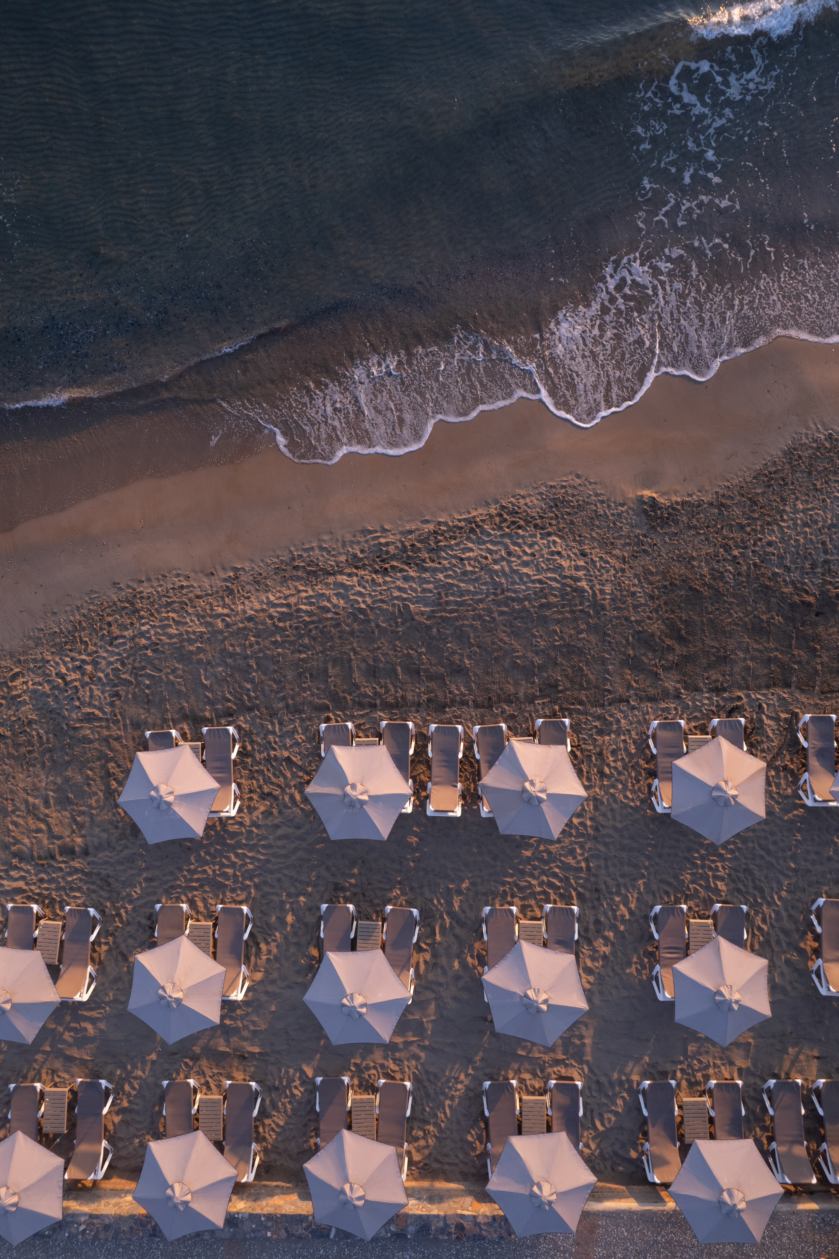 Aerial beach view: rows of grey umbrellas, sun loungers, ocean waves.