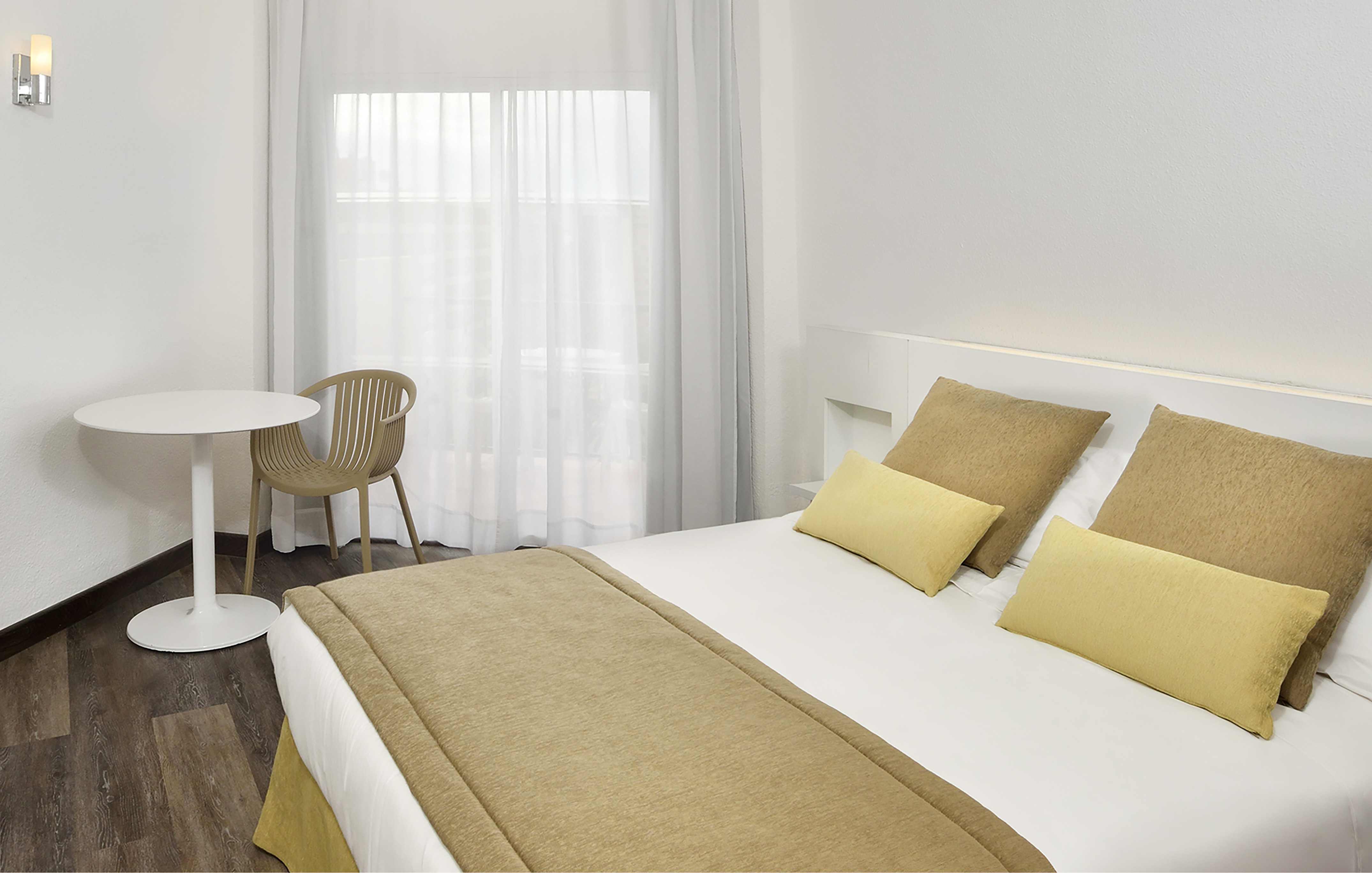 Modern hotel room with white bed, beige linens, table, and window.