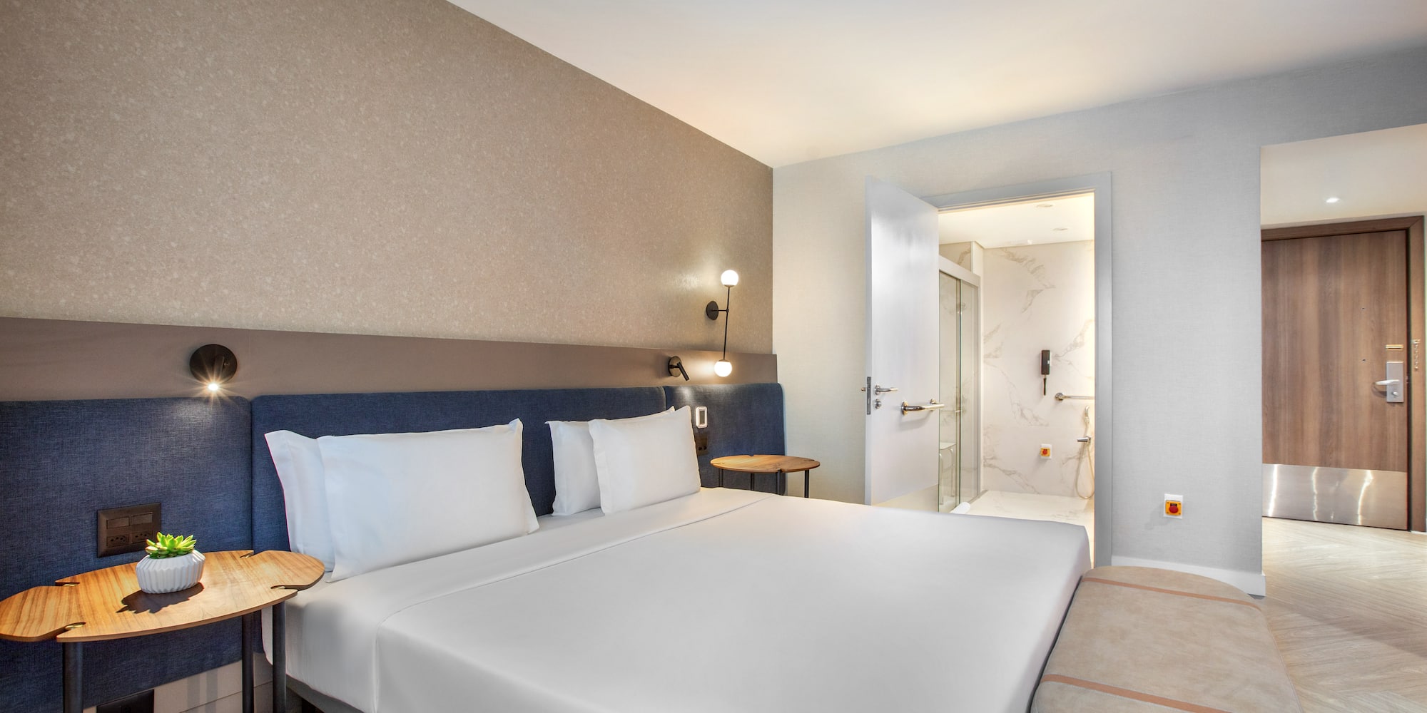 Modern hotel bedroom with white bed, blue headboard, and adjacent bathroom.