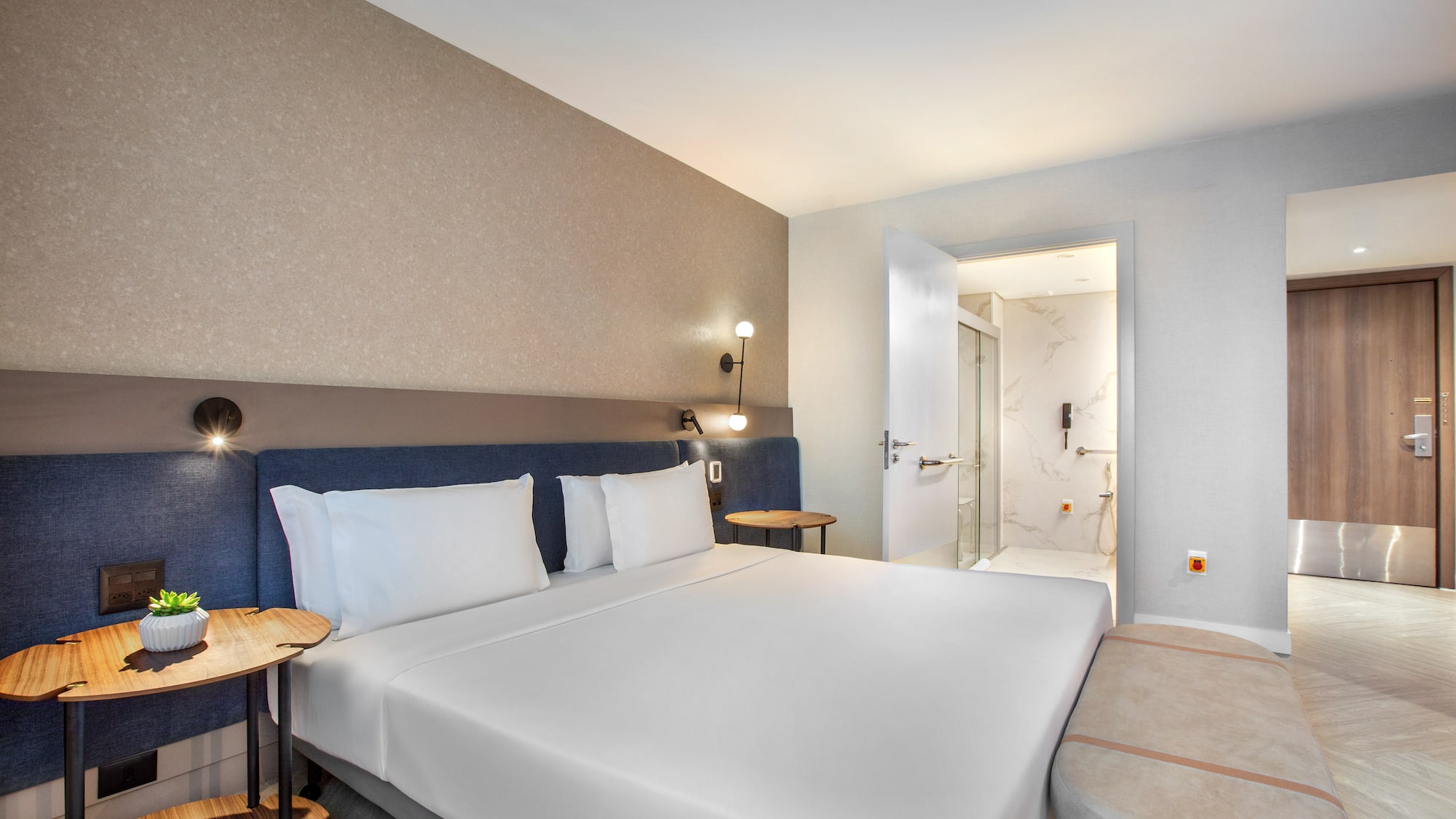 Modern hotel bedroom with white bed, blue headboard, and adjacent bathroom.