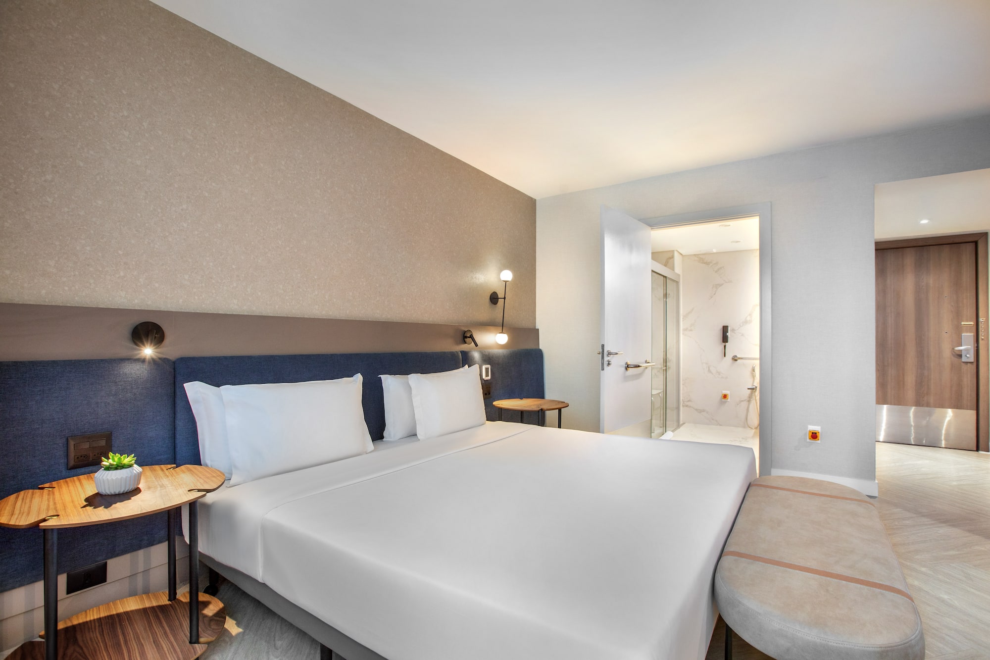 Modern hotel bedroom with white bed, blue headboard, and adjacent bathroom.
