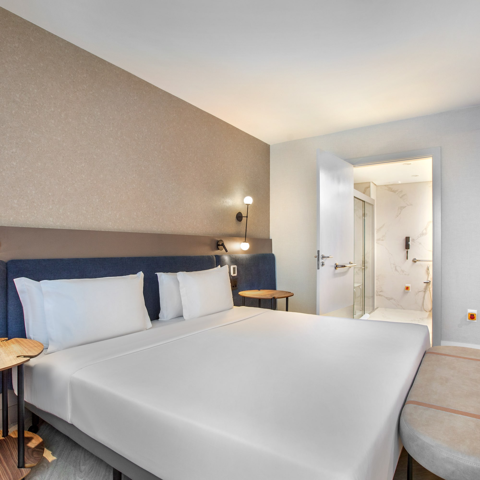 Modern hotel bedroom with white bed, blue headboard, and adjacent bathroom.