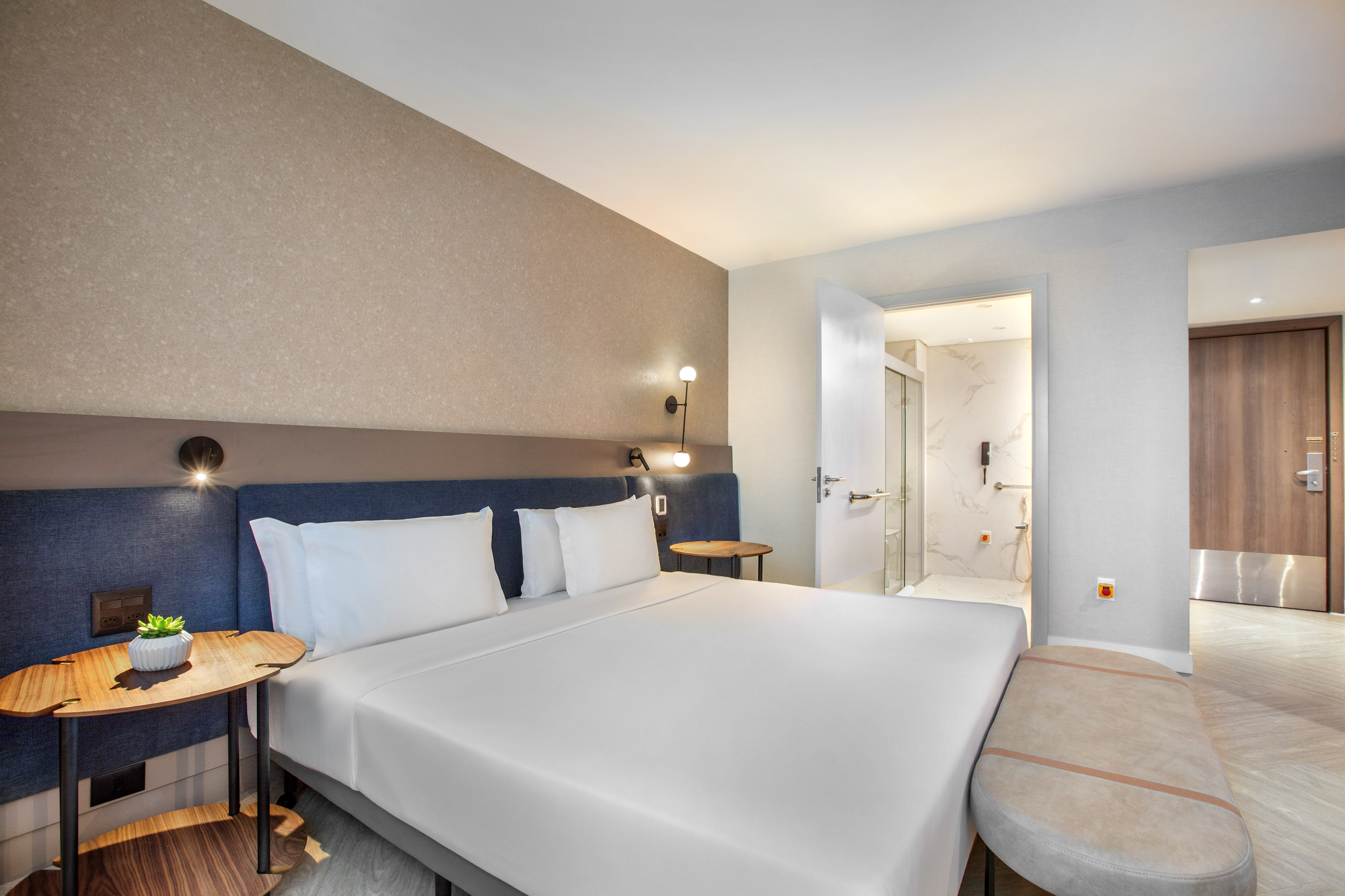 Modern hotel bedroom with white bed, blue headboard, and adjacent bathroom.