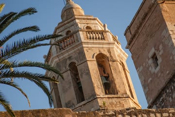 Historic stone bell tower with arched openings, bells, and palm tree under blue sky.