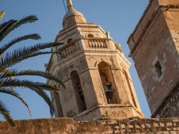 Historic stone bell tower with arched openings, bells, and palm tree under blue sky.