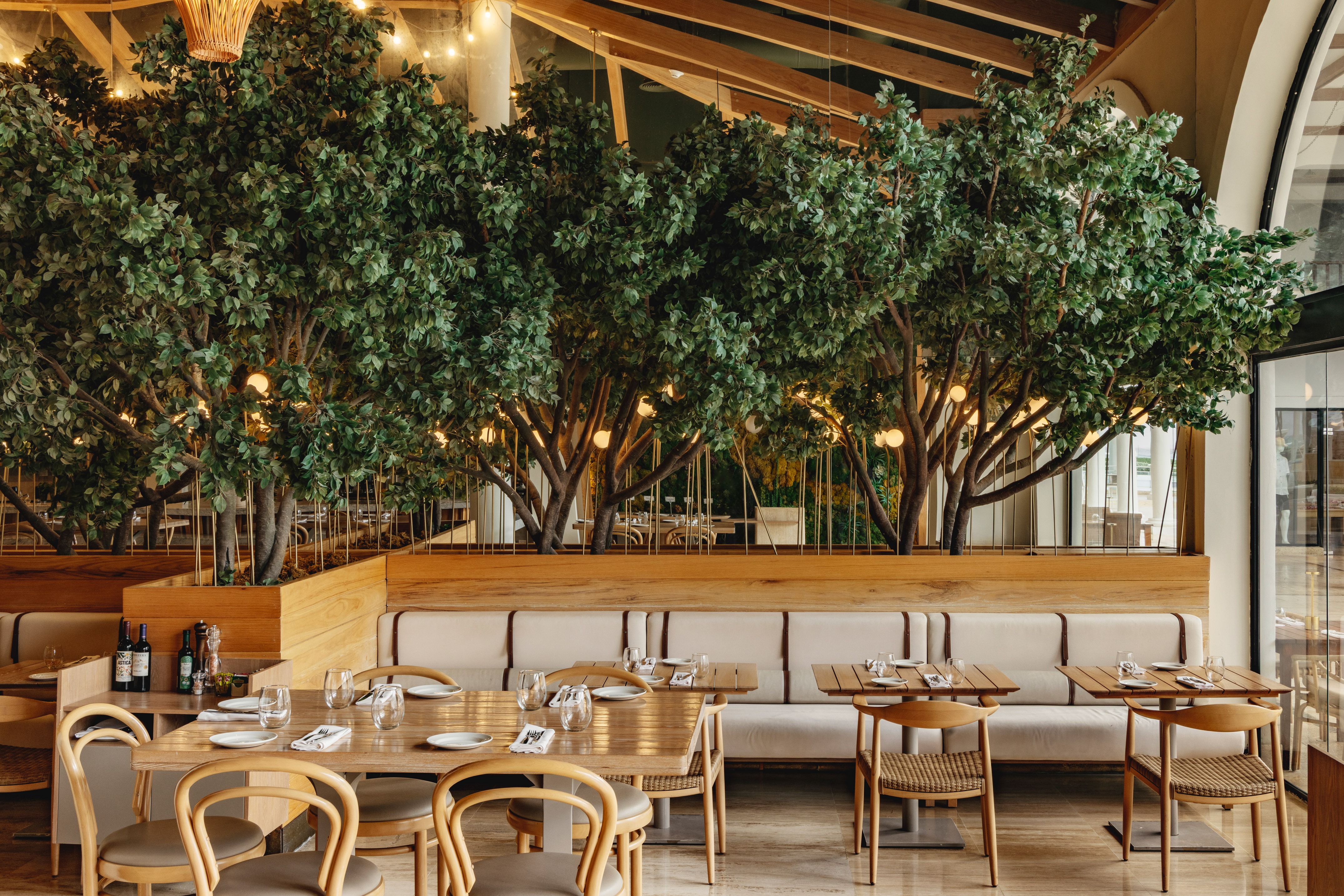 Elegant restaurant dining room with indoor trees, wooden tables, and booths.