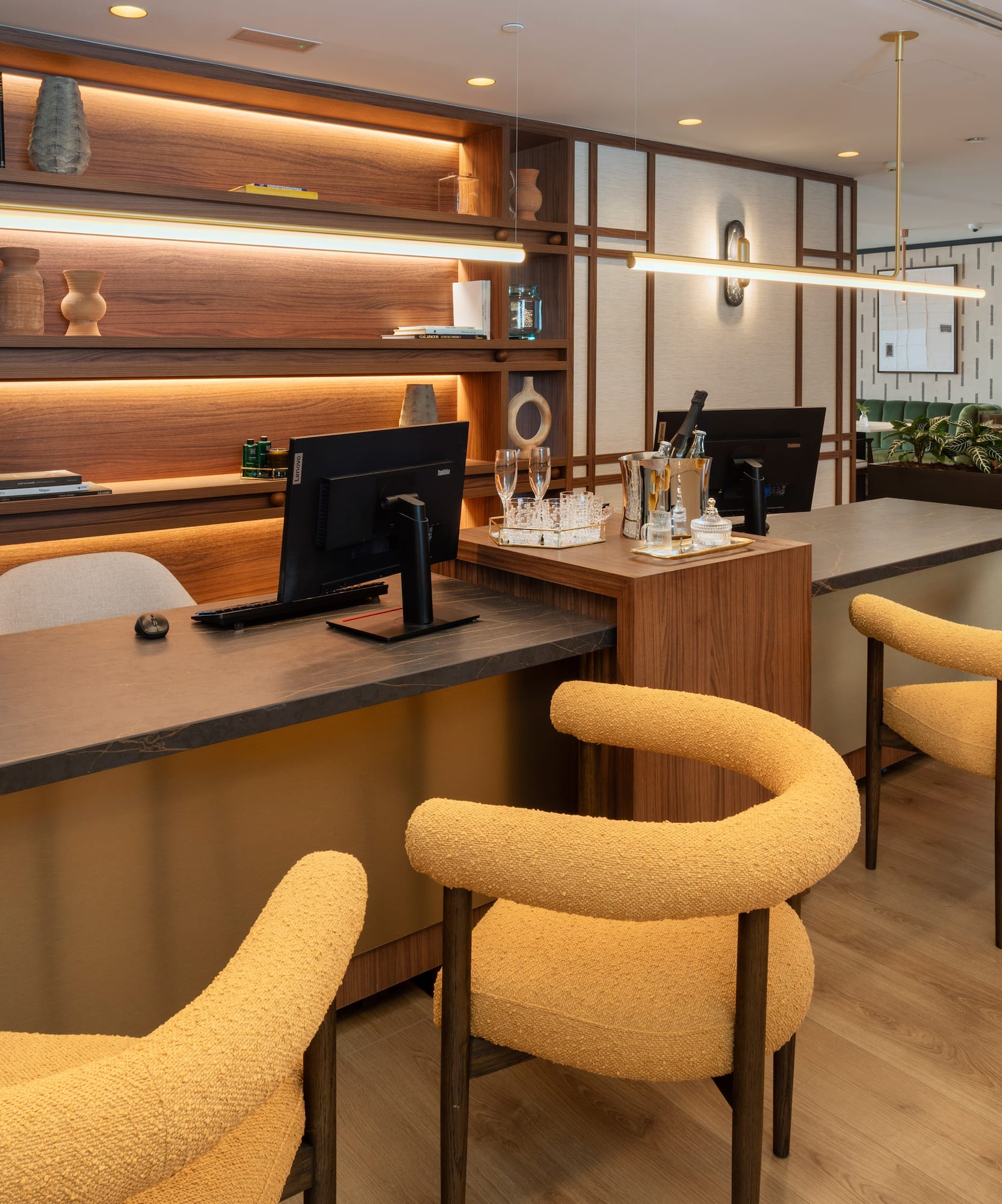 Modern hotel reception desk with yellow upholstered chairs and backlit wooden shelves.