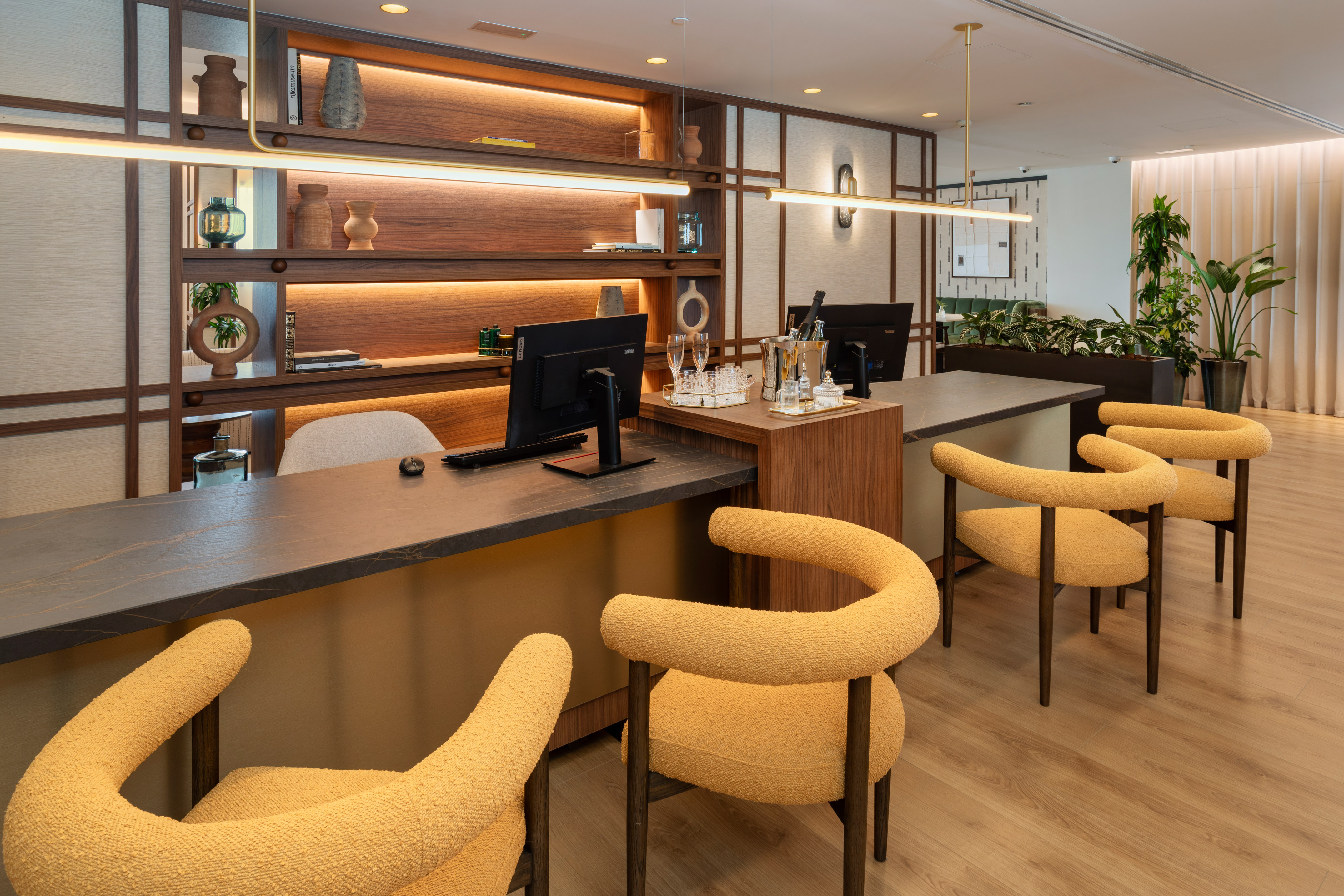 Modern hotel reception desk with yellow upholstered chairs and backlit wooden shelves.