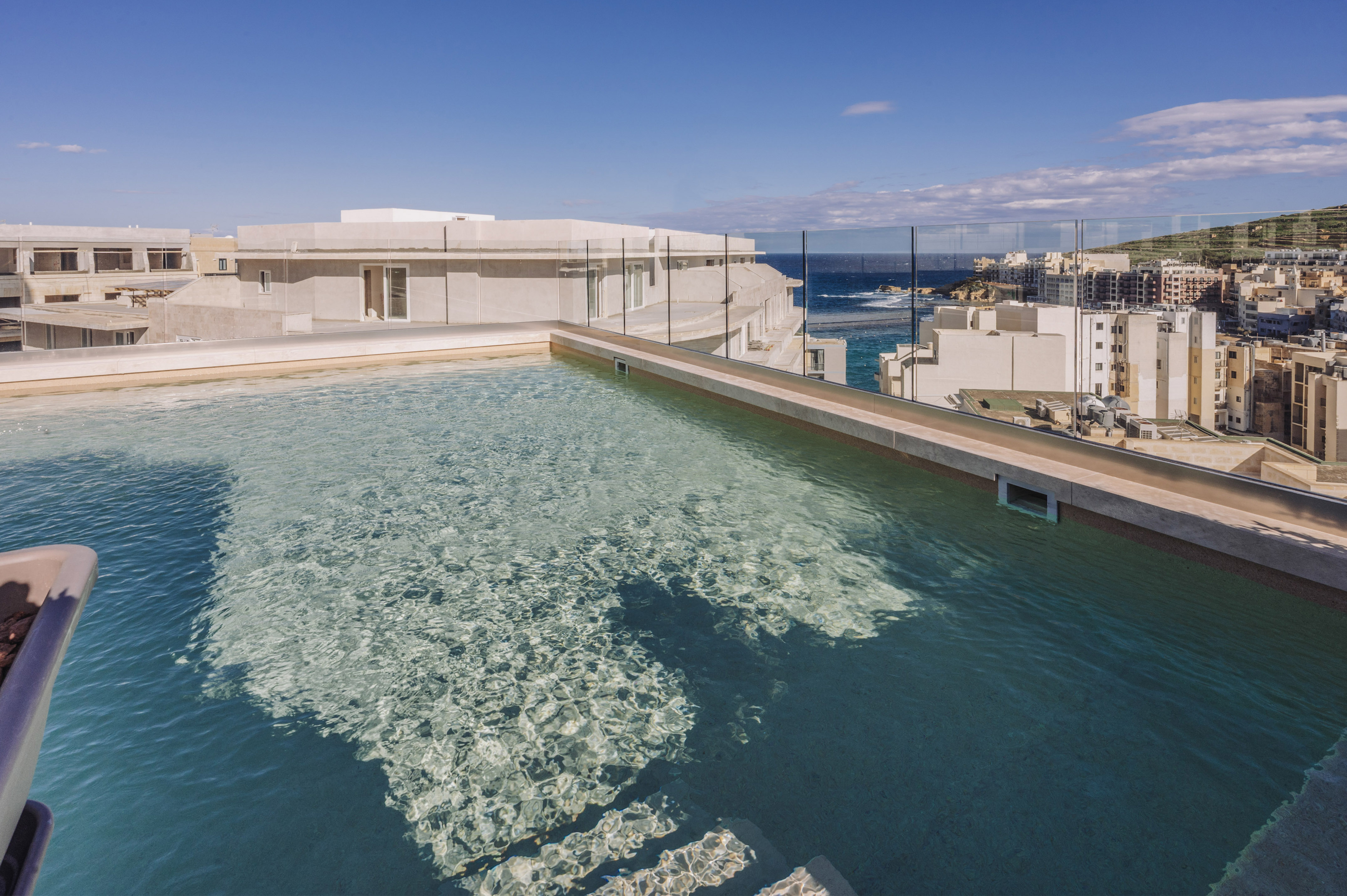 Clear rooftop pool overlooking a sunny coastal city and blue sea.