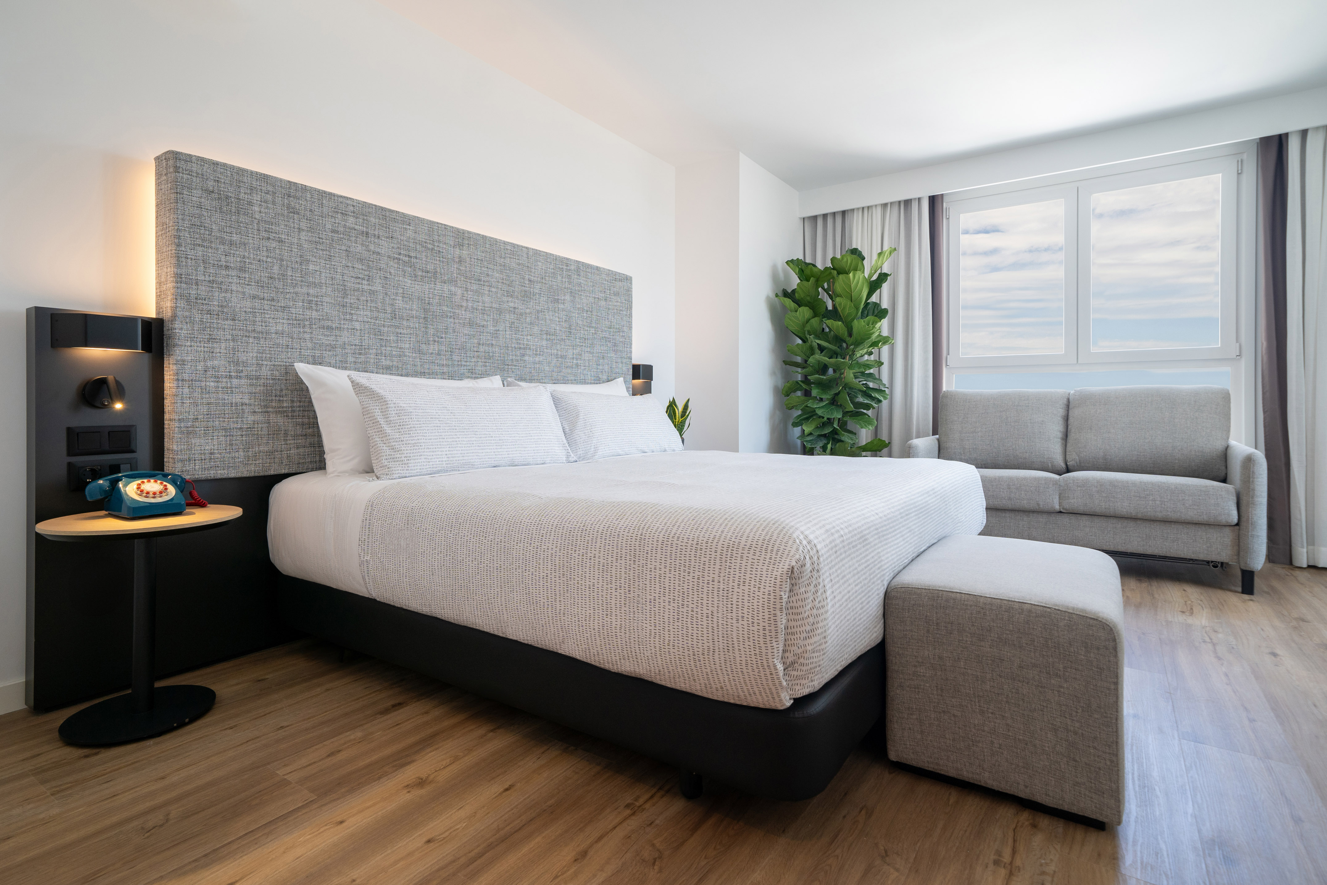 Modern hotel bedroom with king bed, gray upholstered headboard, sofa, window view.