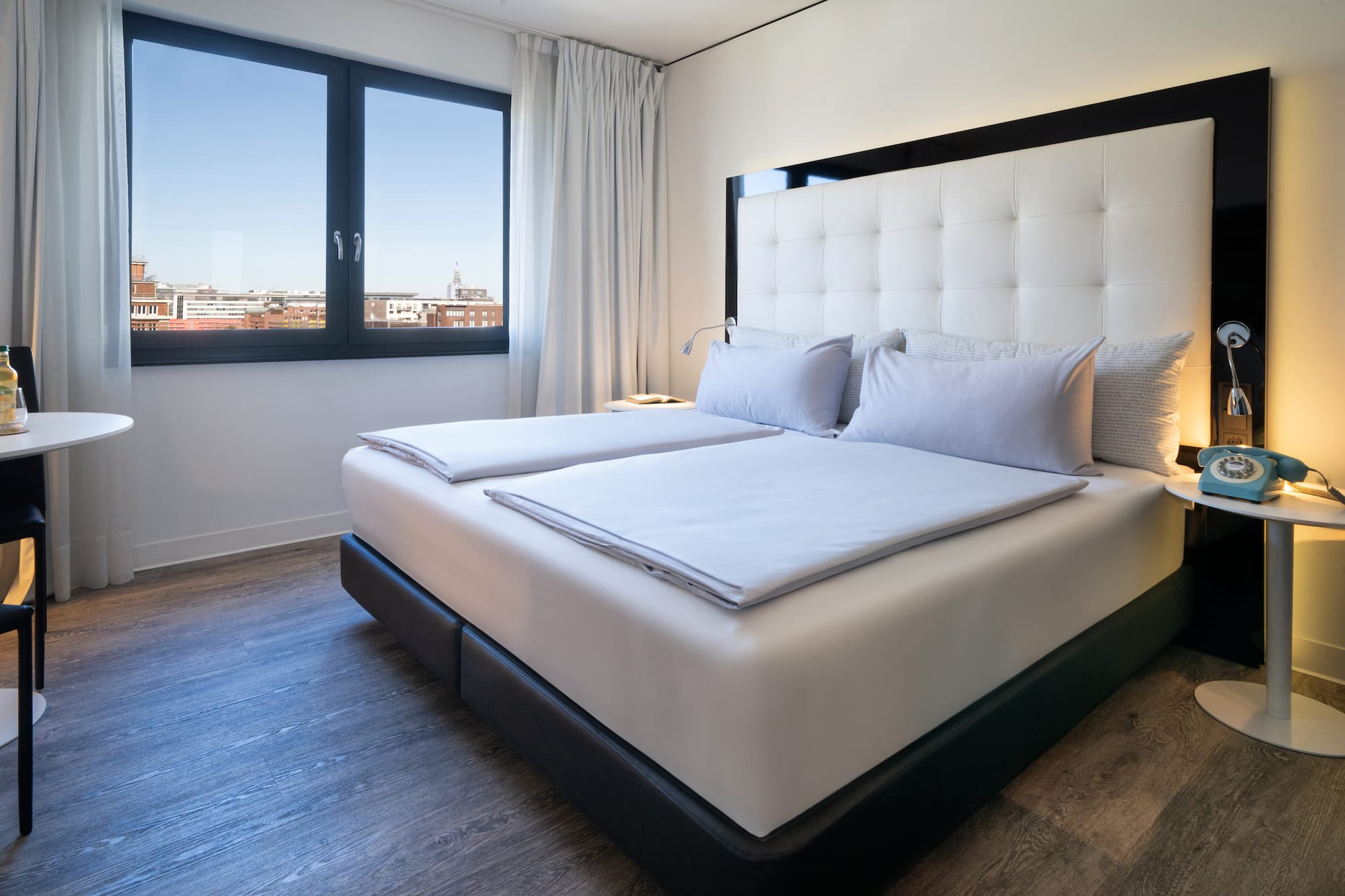 Modern hotel bedroom with city view, white tufted bed, and retro blue phone.