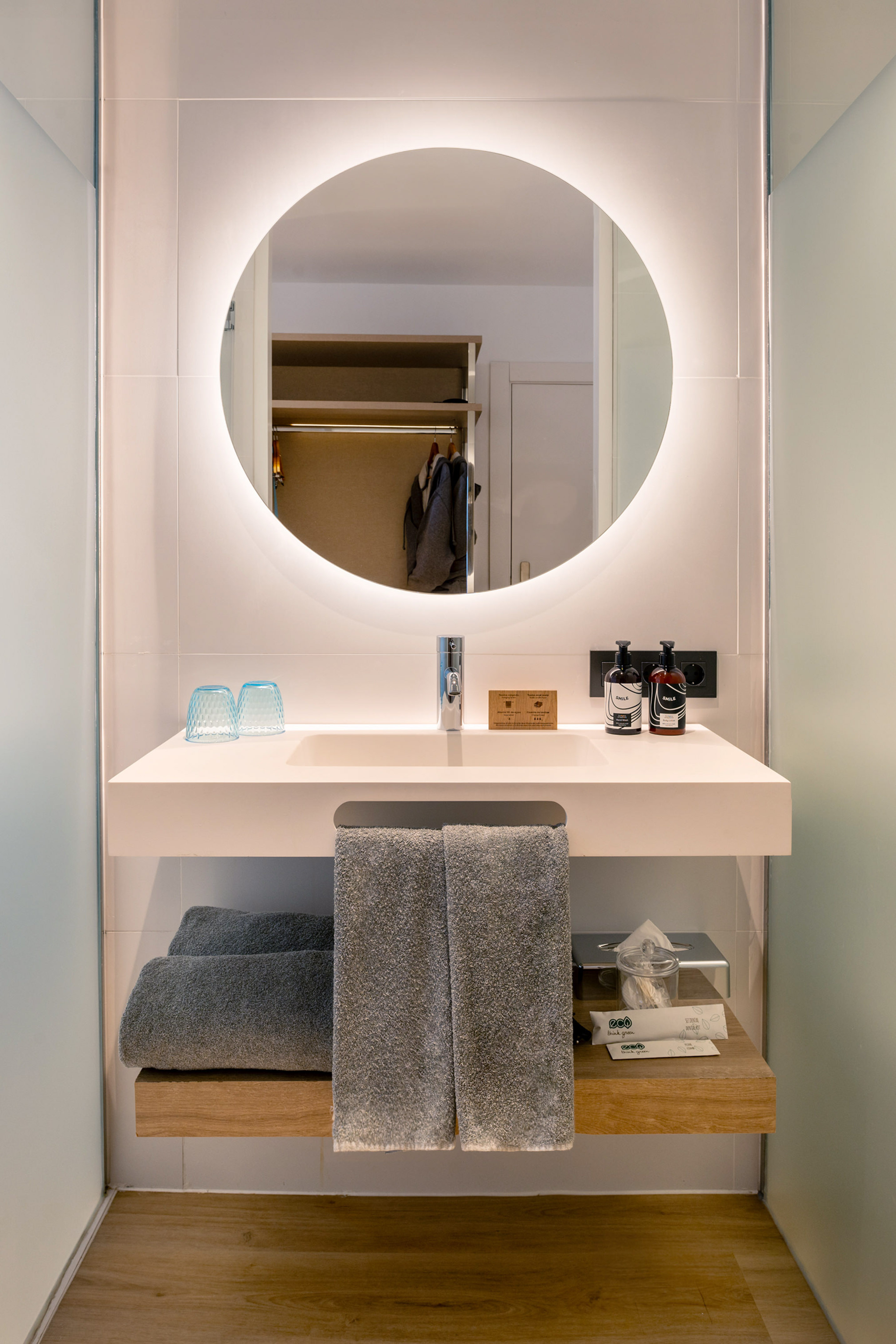 Modern bathroom vanity with illuminated mirror, white sink, grey towels, and toiletries.