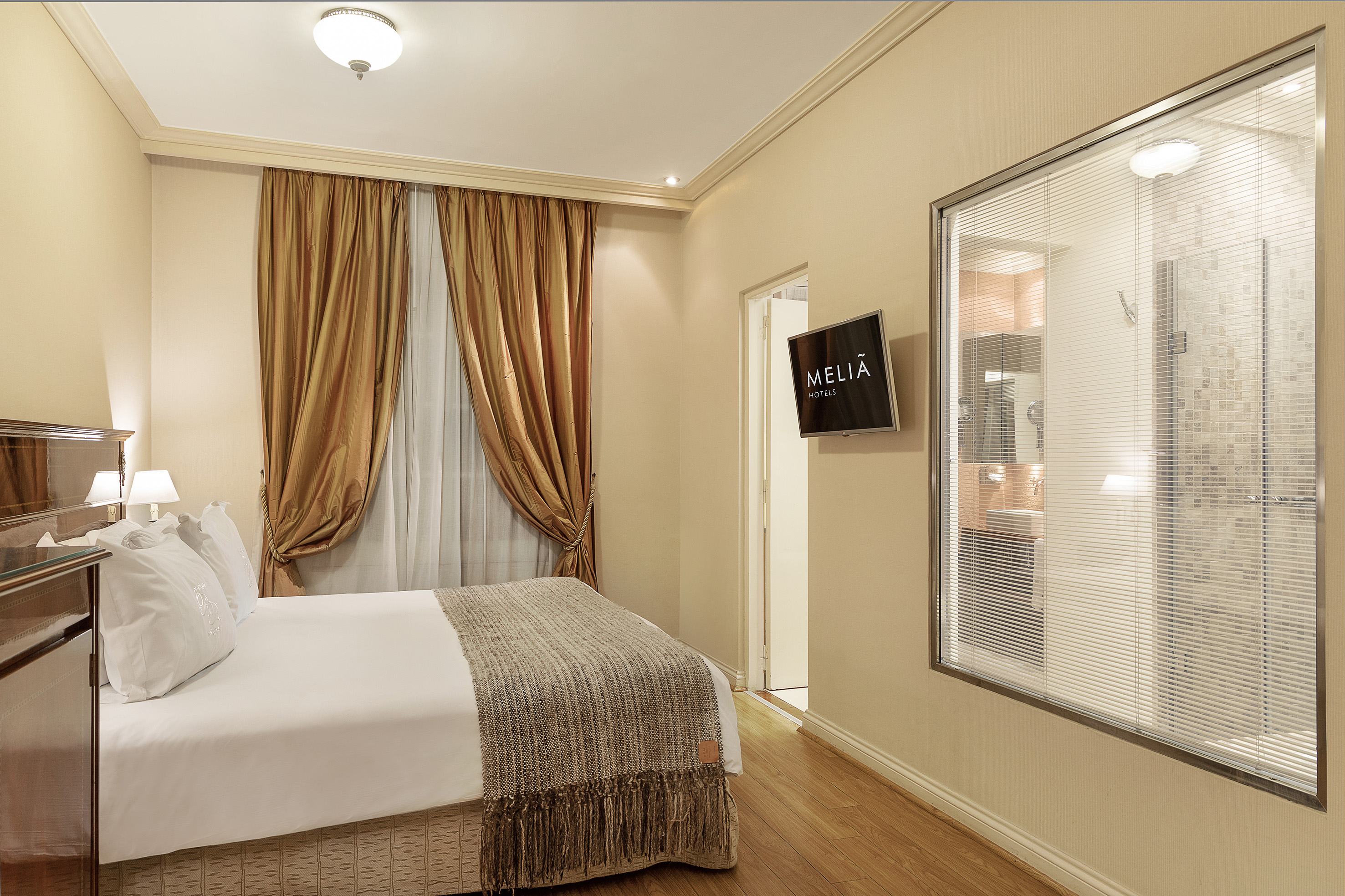 Meliã Hotels guest bedroom with bed, gold curtains, wood floor, and glass shower.