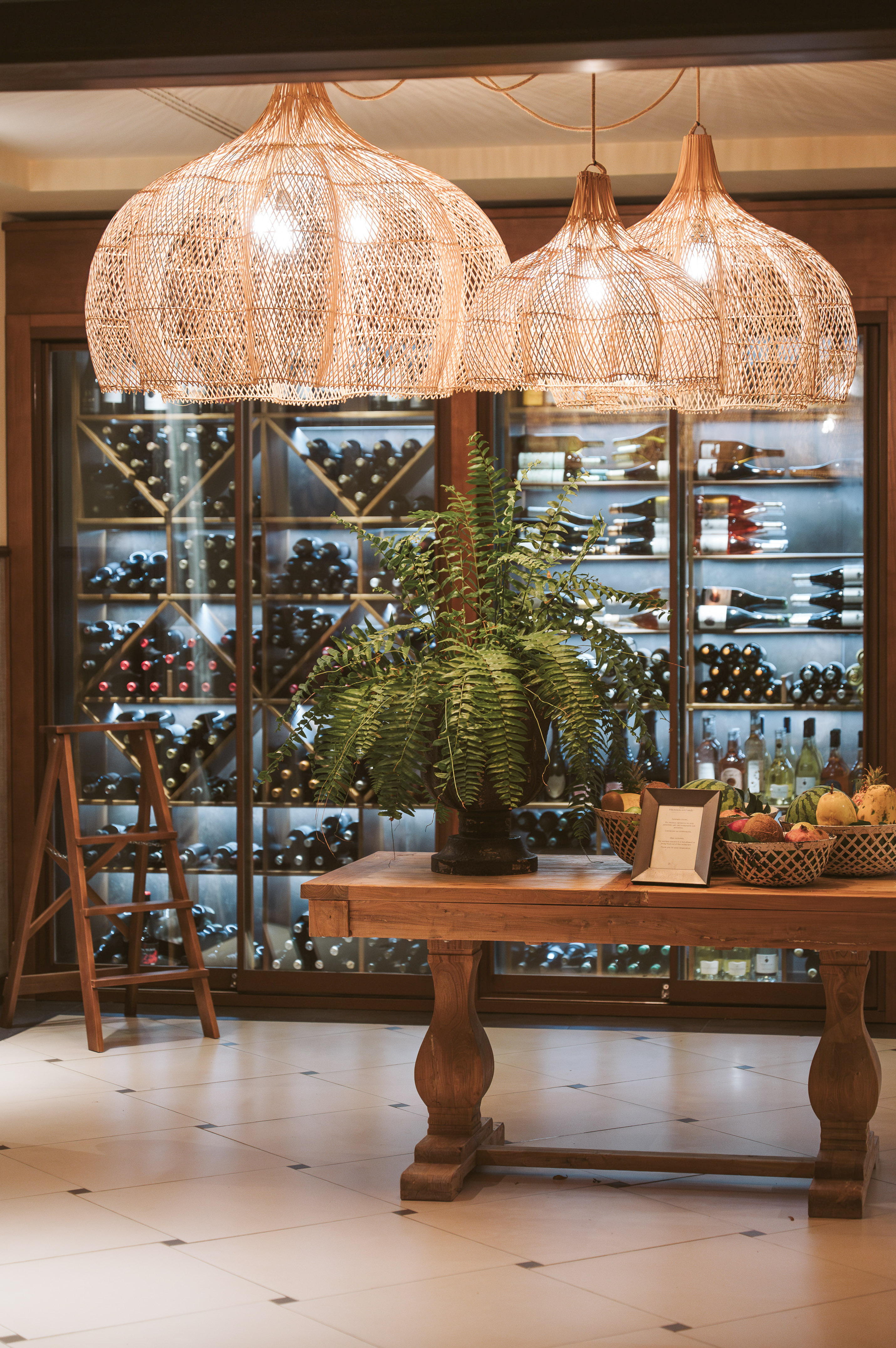 Wine cellar with wooden table, potted fern, fruit baskets, rattan lights.