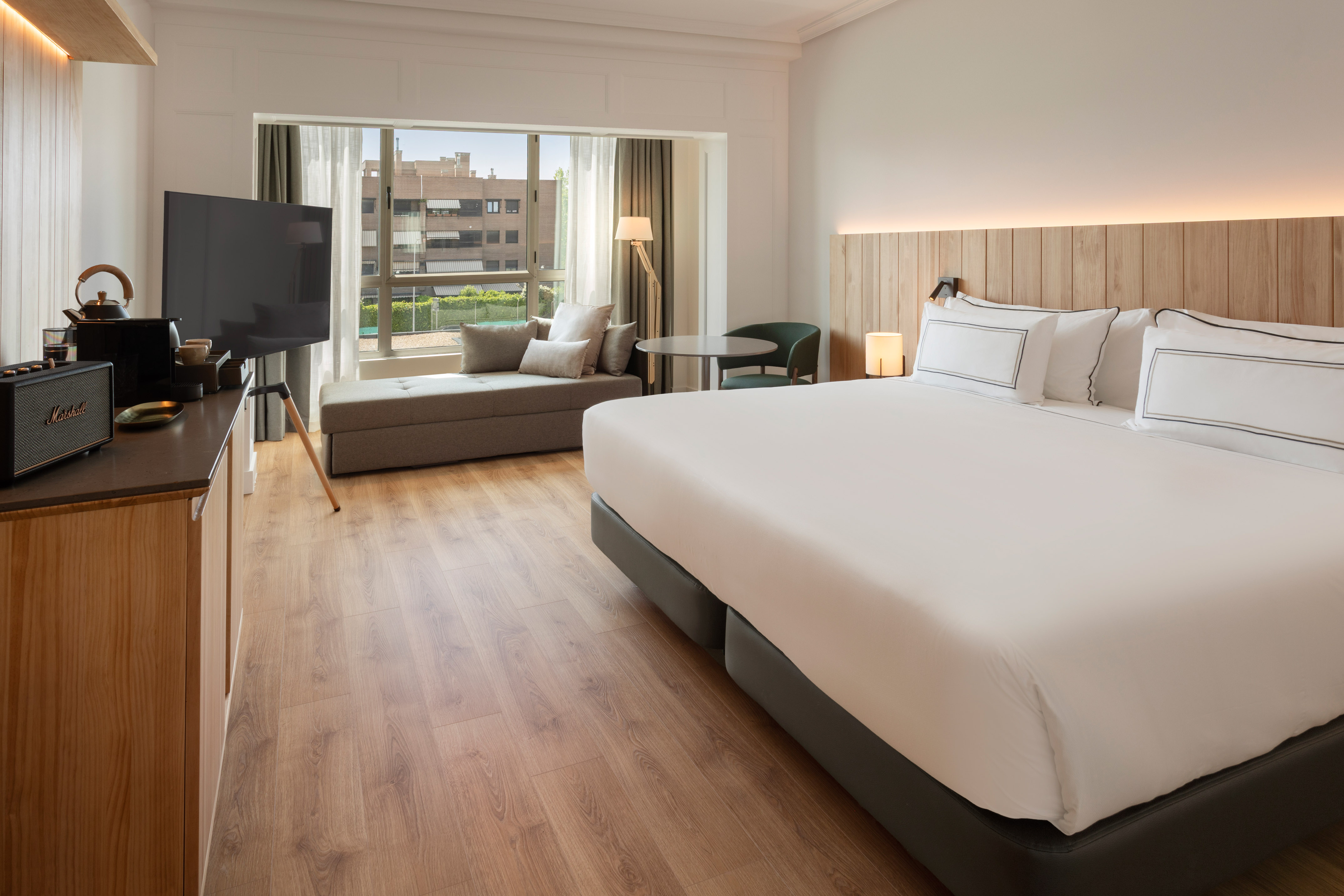 Modern hotel room with king bed, wooden headboard, window view, daybed, and TV.