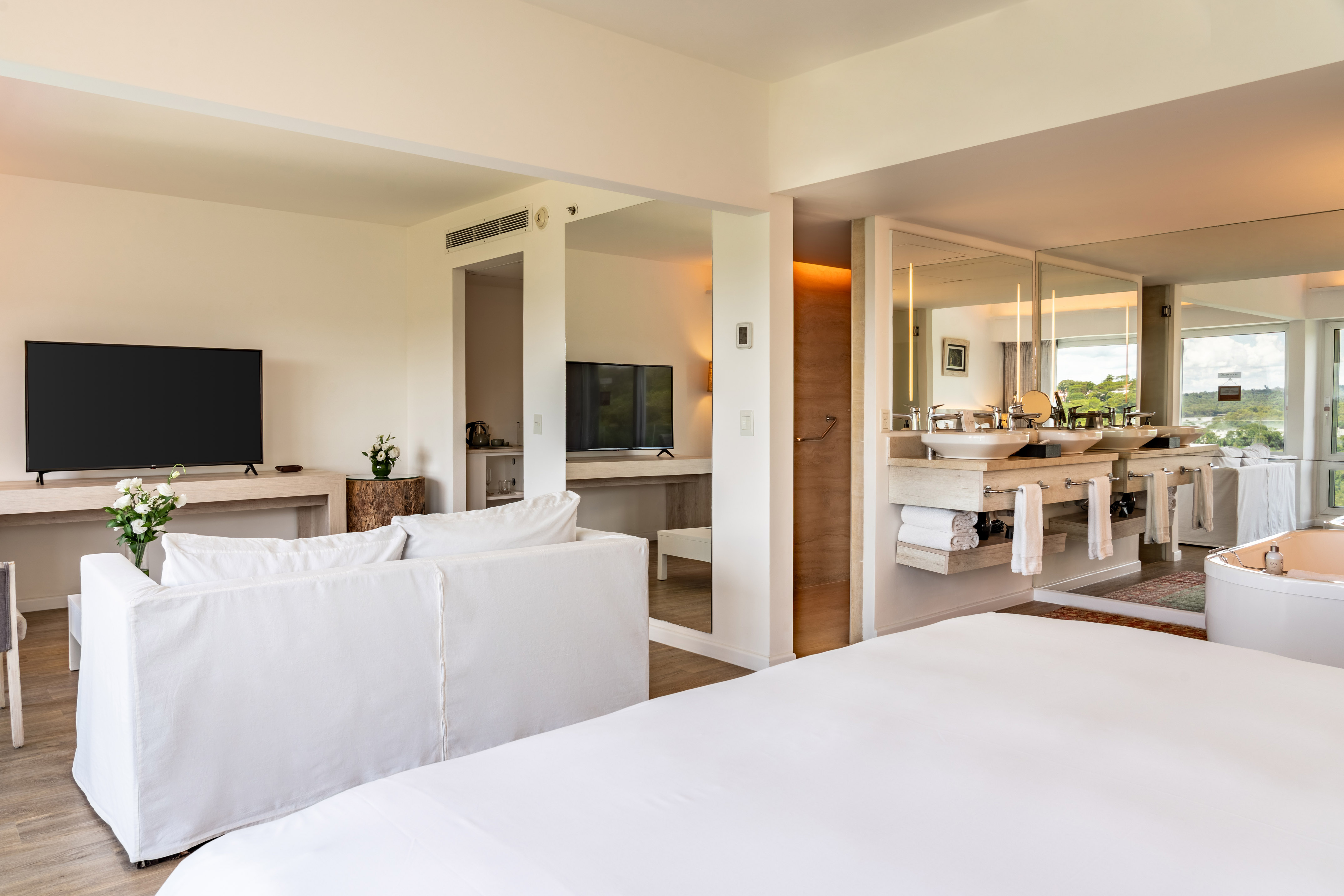 Modern hotel suite with bed, sofa, double sink bathroom, and freestanding tub.