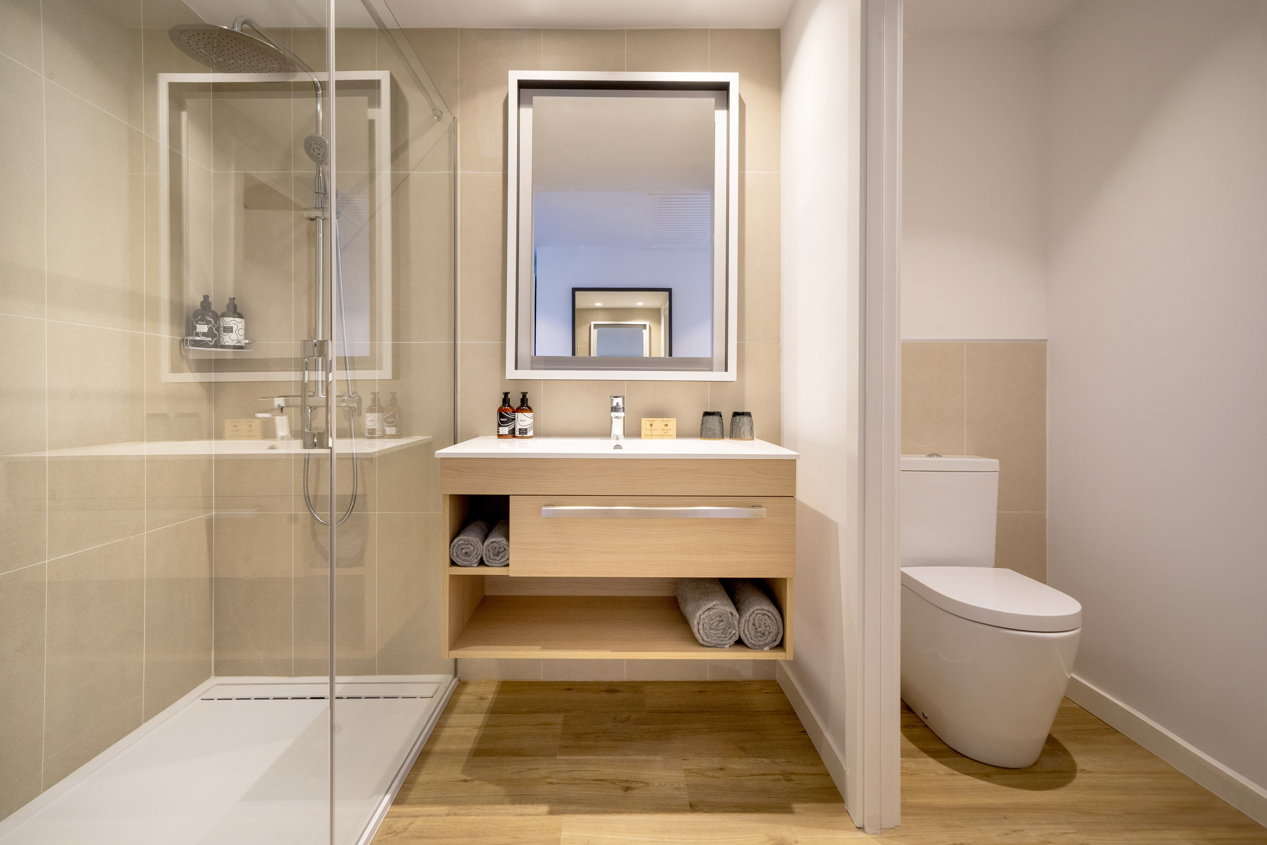 Sleek modern hotel bathroom with glass shower, wood vanity, mirror, and toilet.