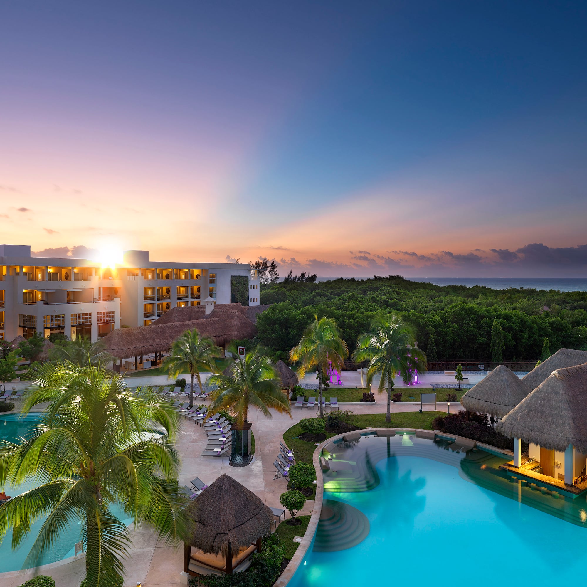 a pool with palm trees and a building with a beach and a sunset