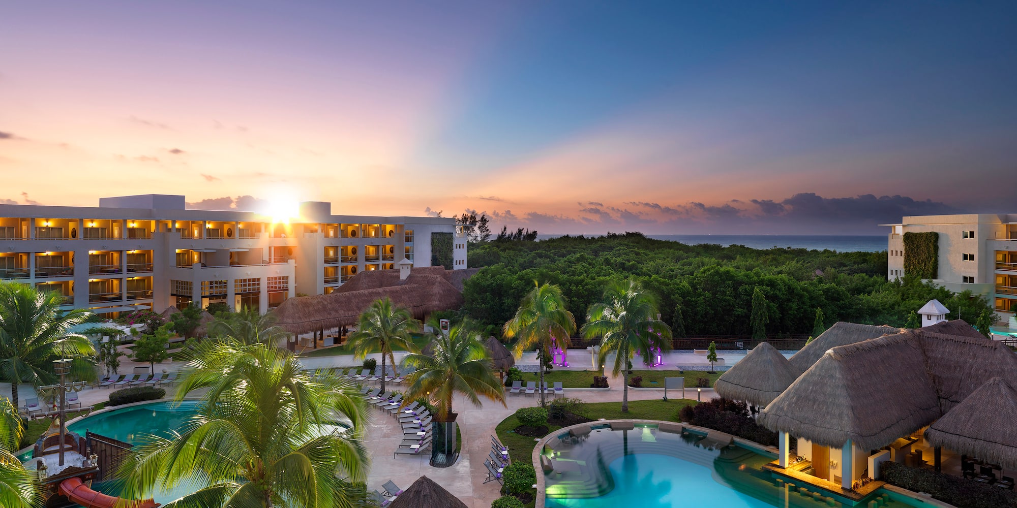 a pool with palm trees and a building with a beach and a sunset