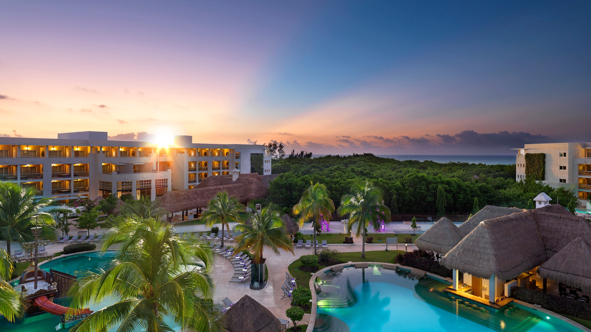 a pool with palm trees and a building with a beach and a sunset