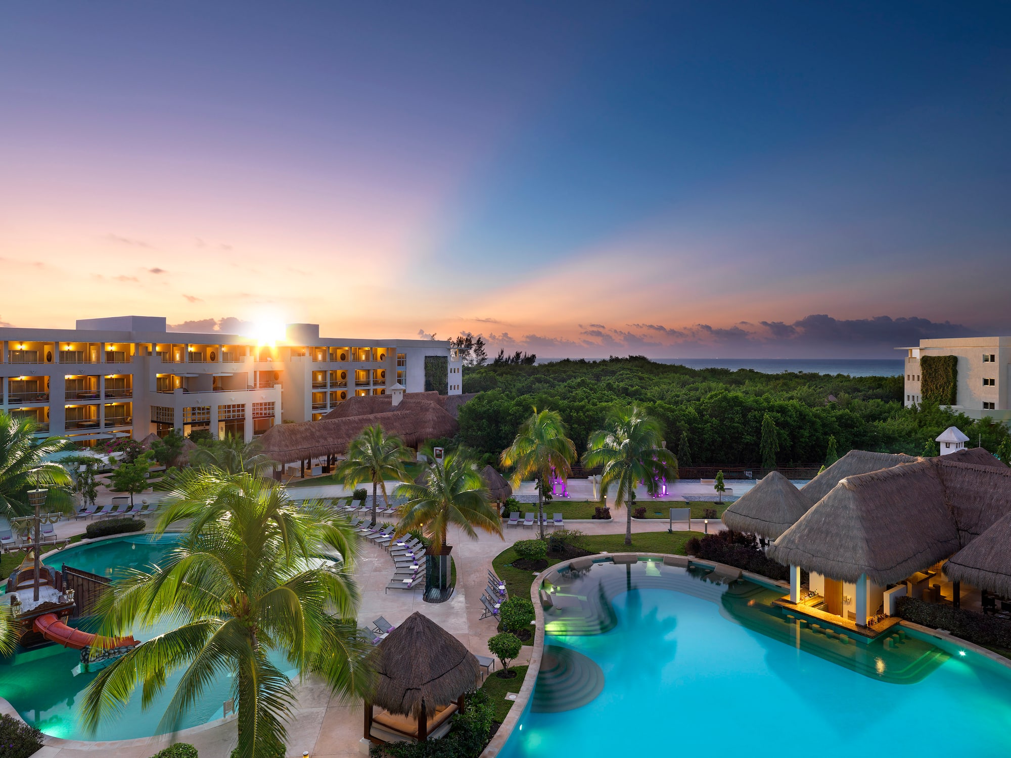 a pool with palm trees and a building with a beach and a sunset