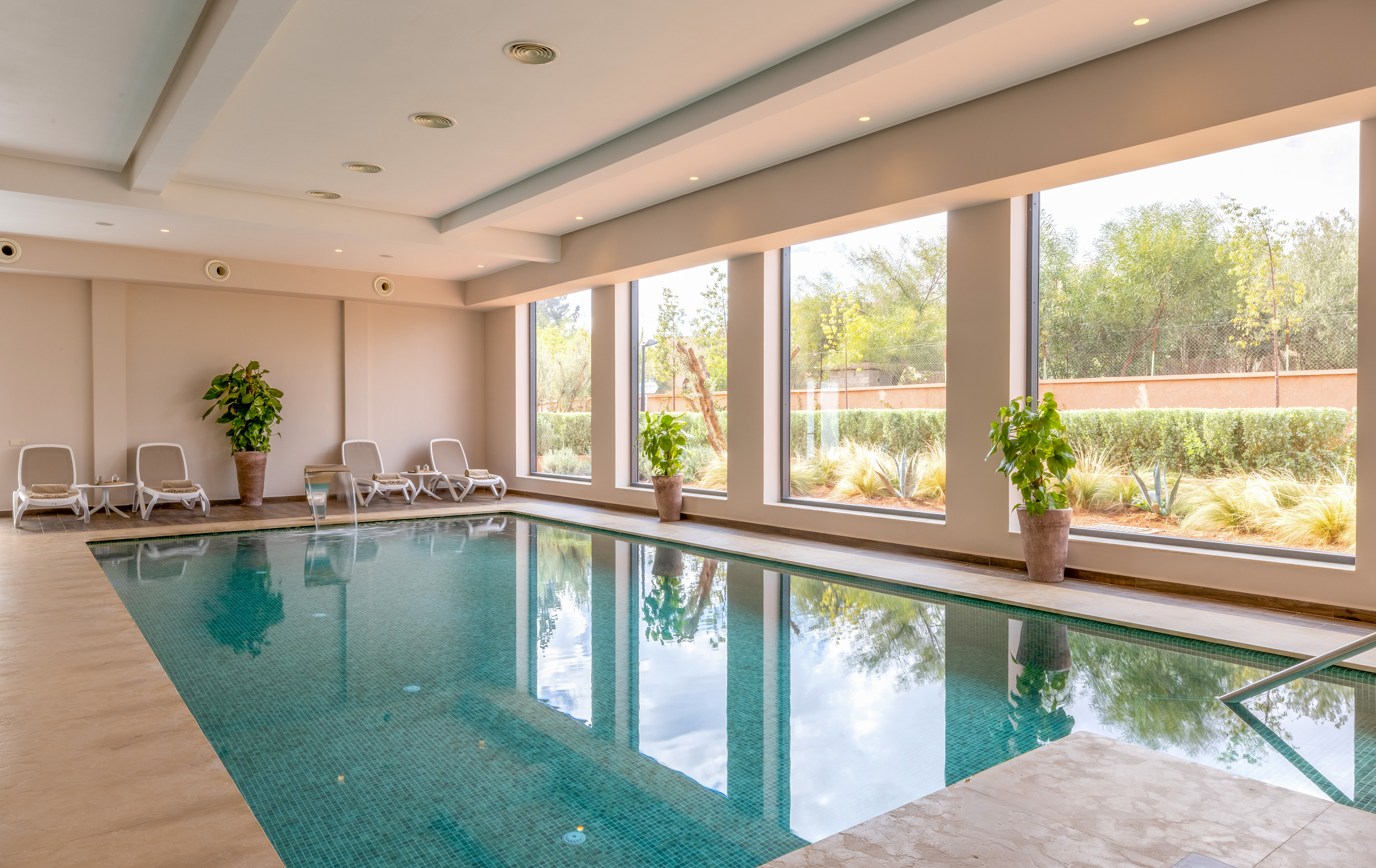 Modern indoor hotel swimming pool with lounge chairs, large windows, garden view.
