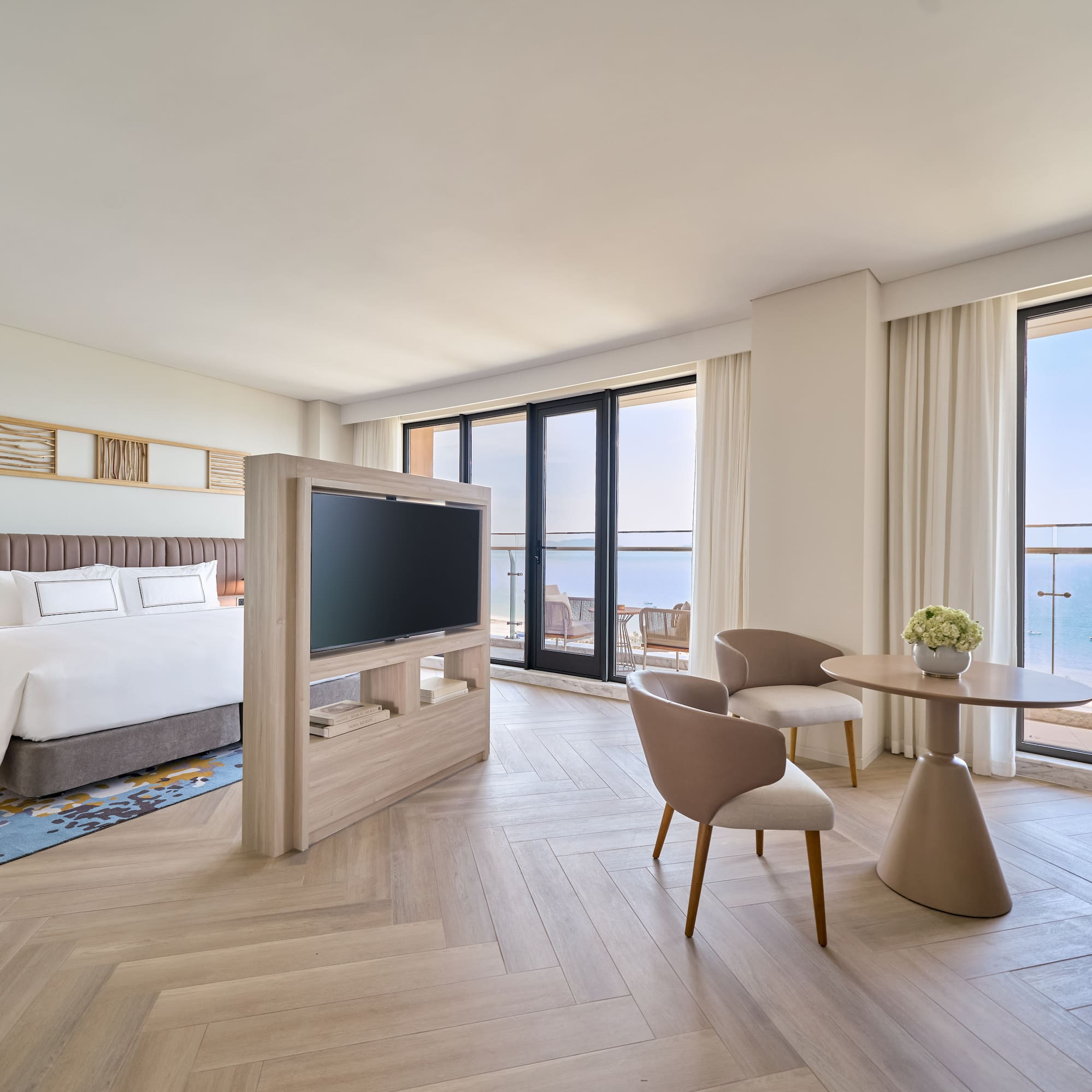 Modern ocean view hotel suite with bed, TV, and seating by the balcony.