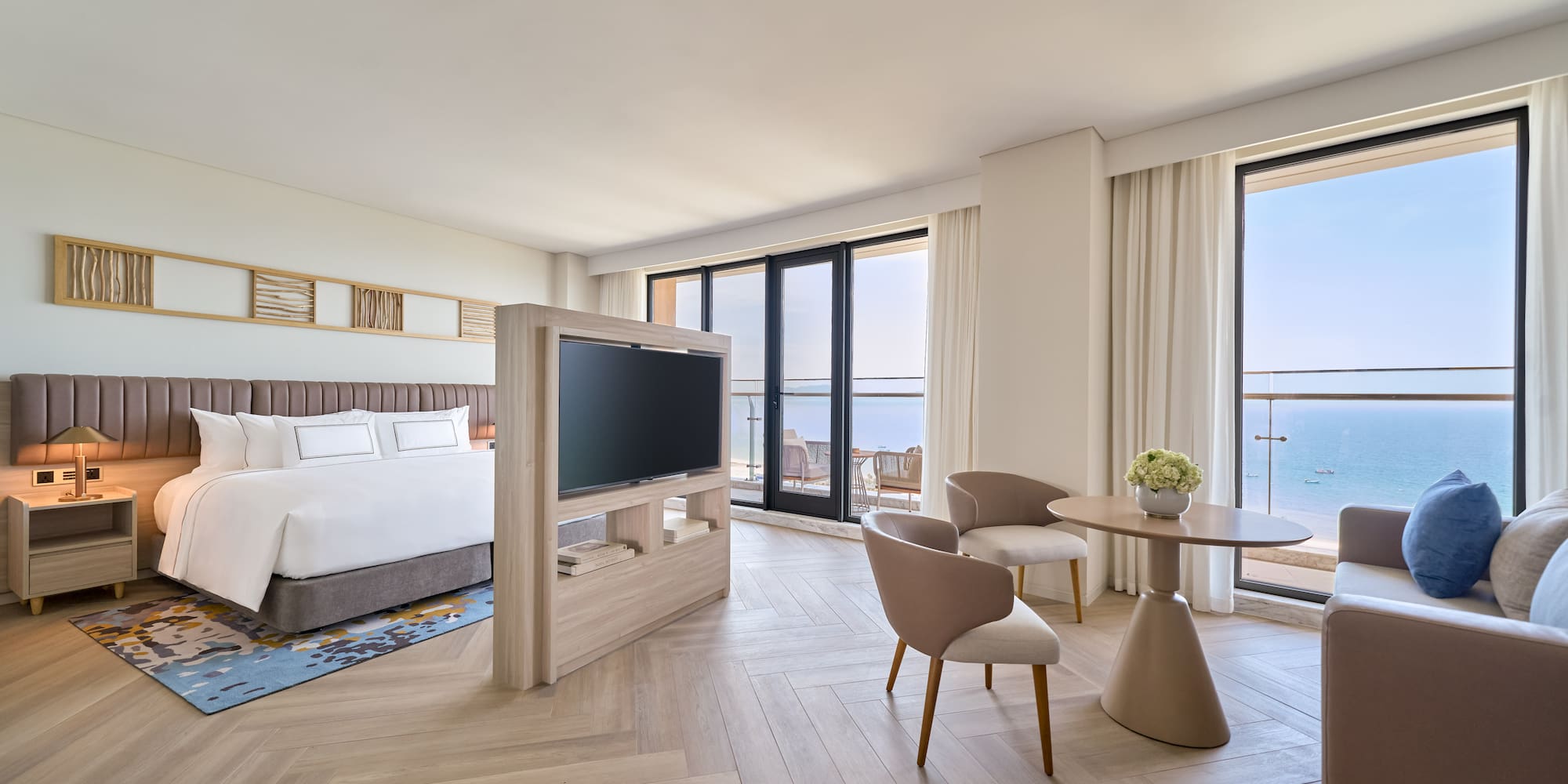 Modern hotel suite with ocean view, large bed, TV, and seating area.