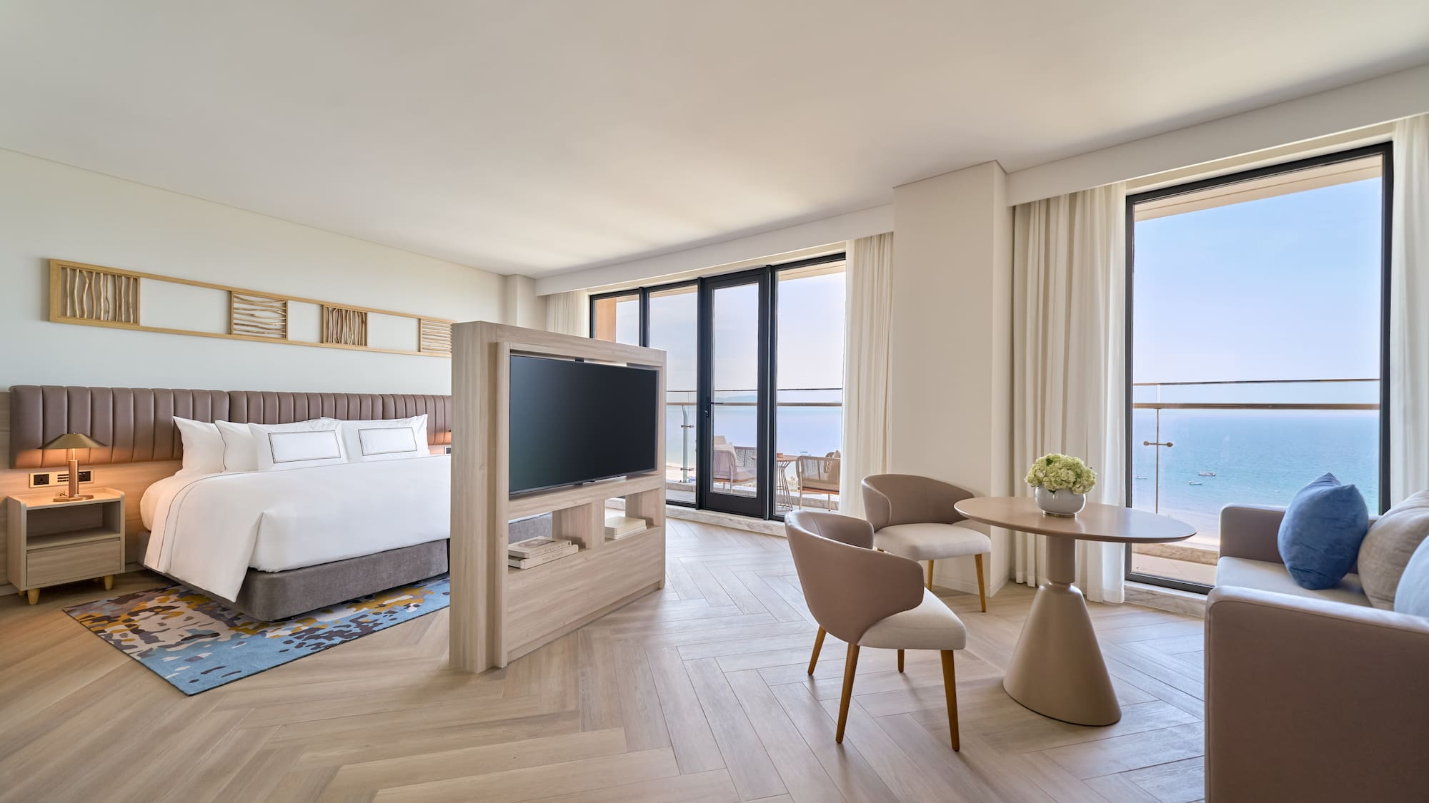 Modern ocean view hotel suite with bed, TV, and seating by the balcony.