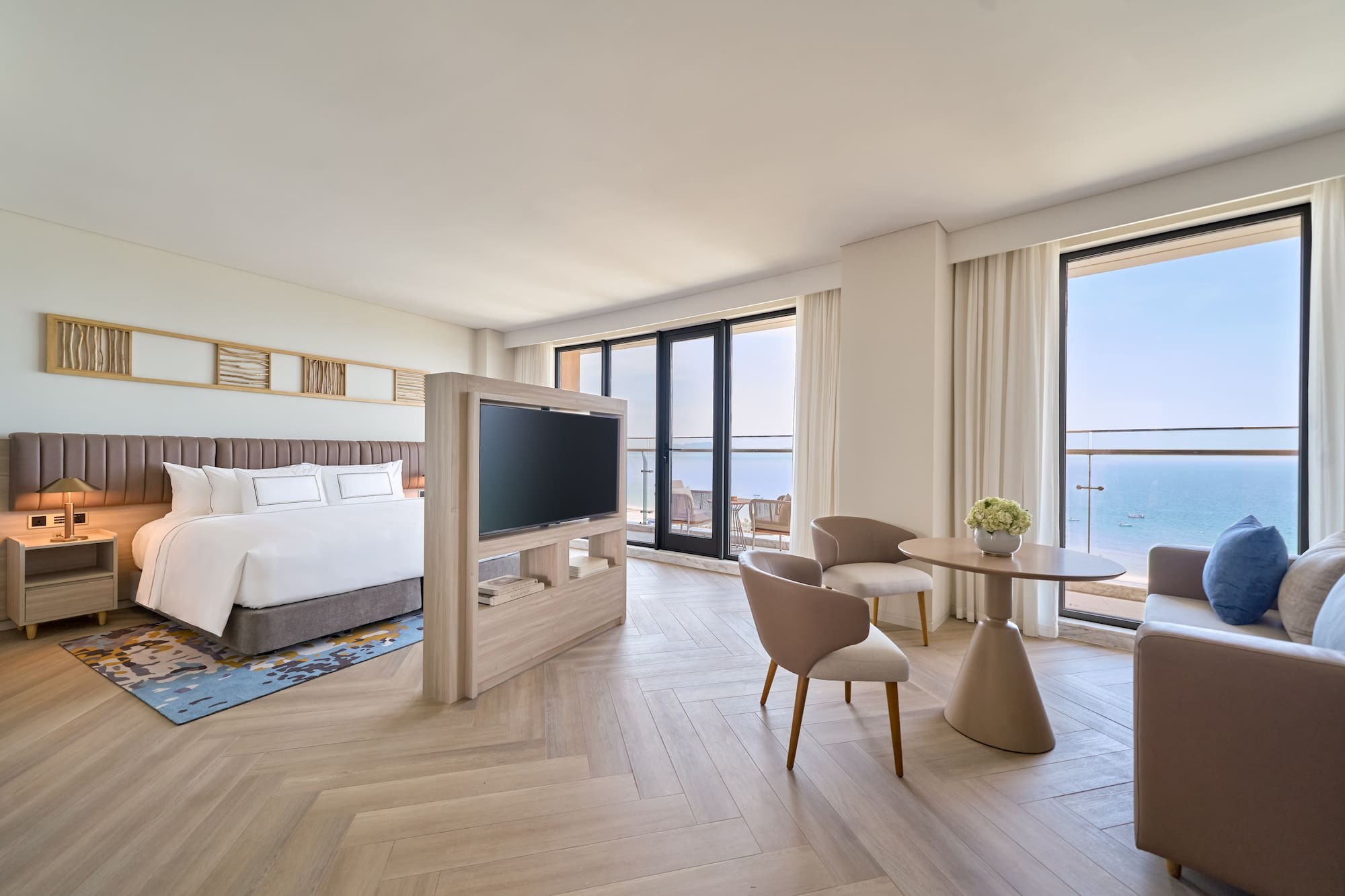 Modern ocean view hotel suite with bed, TV, and seating by the balcony.