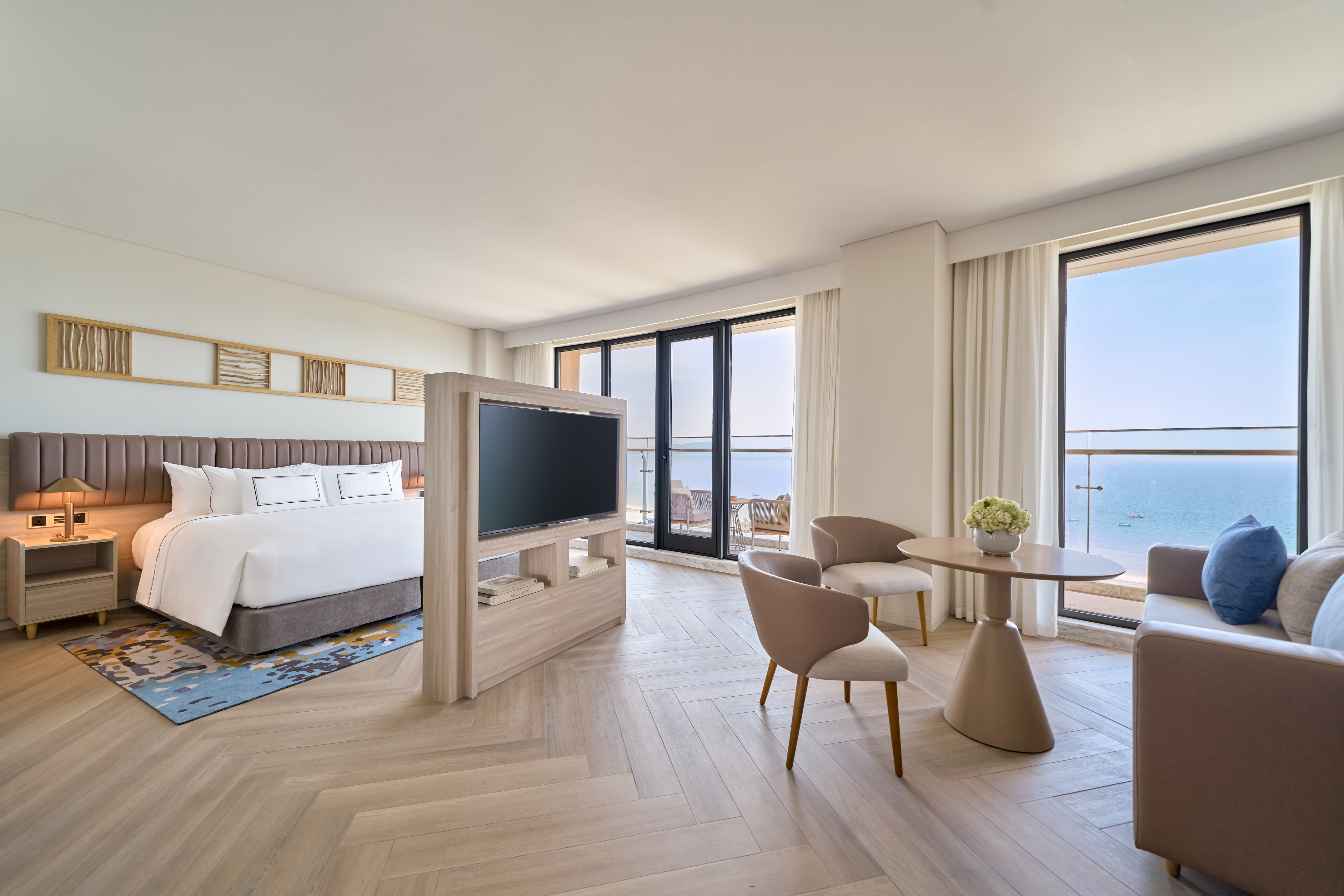 Modern ocean view hotel suite with bed, TV, and seating by the balcony.