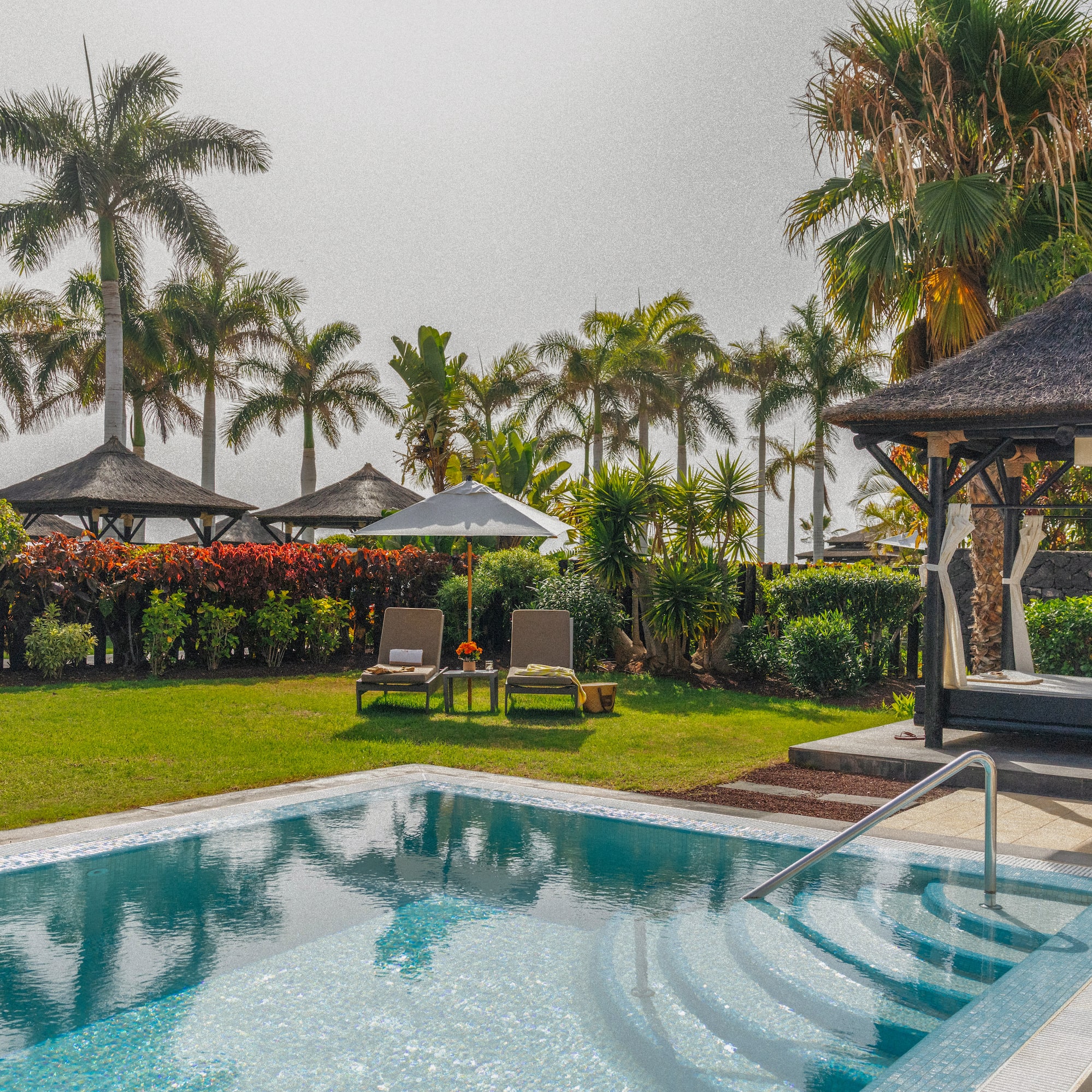 Private swimming pool, palm trees, sun loungers, and thatched gazebo in a garden.