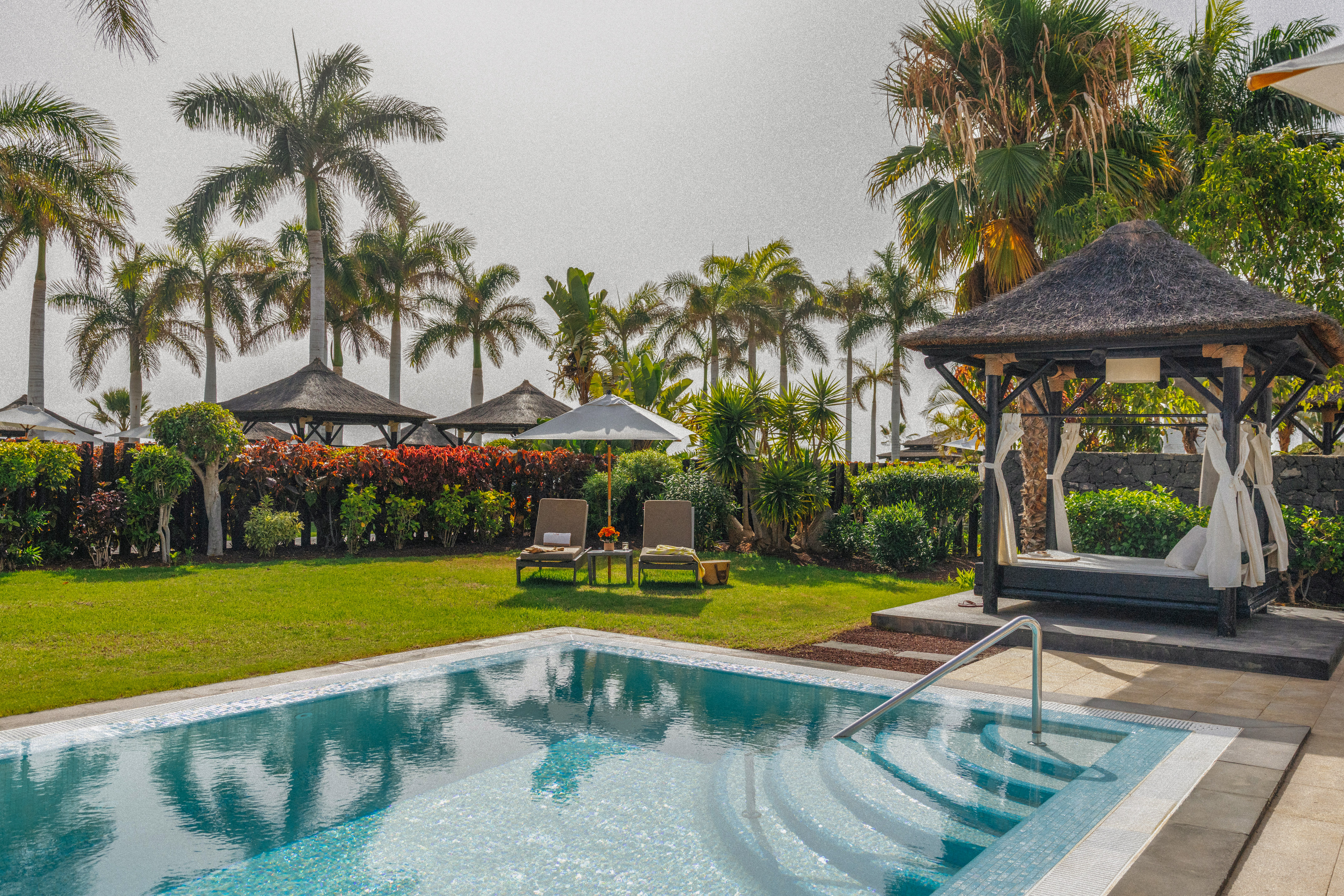 Private resort pool area with palm trees, sunbeds, and thatched cabana.