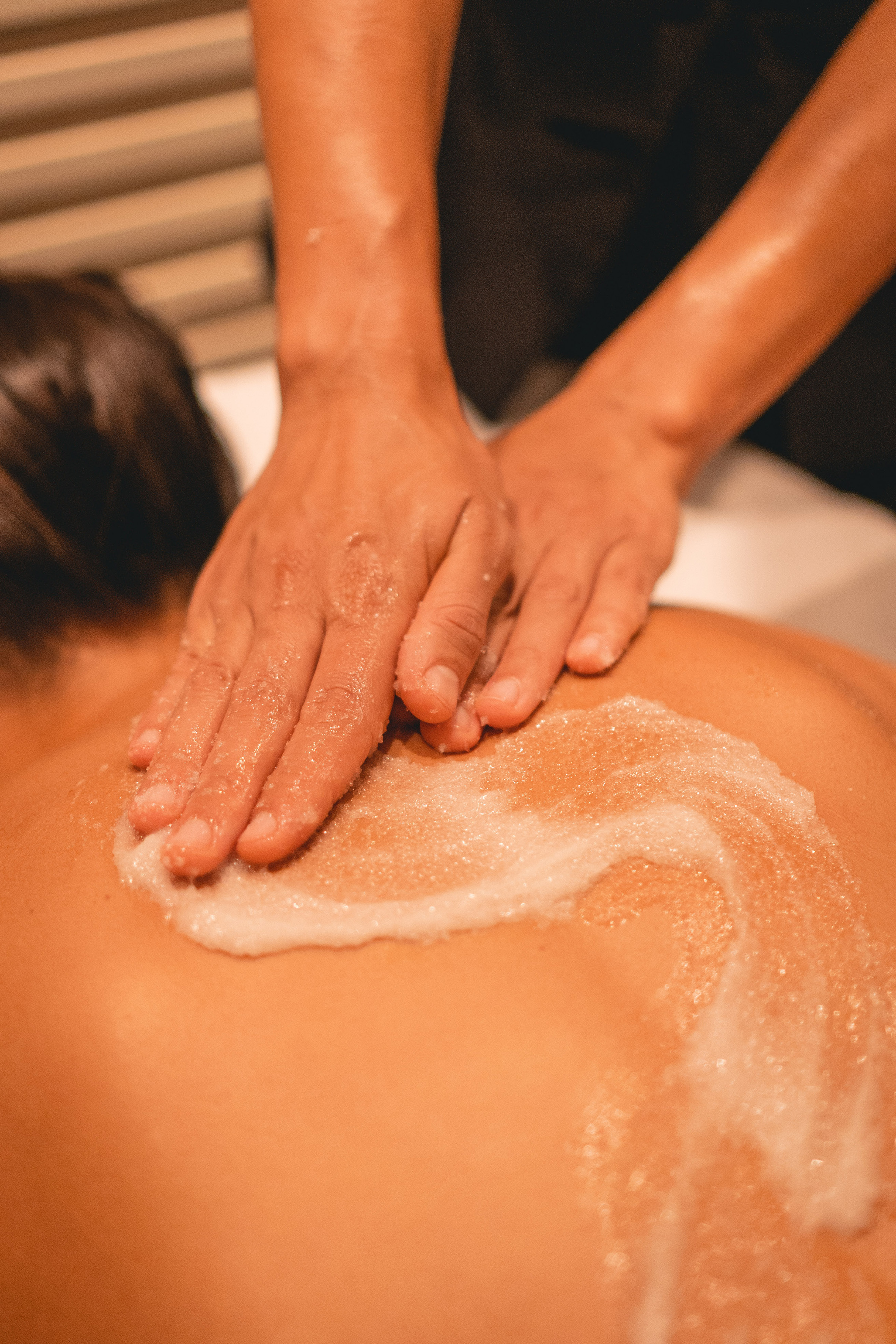 Hands applying exfoliating body scrub to a person's back at a spa.
