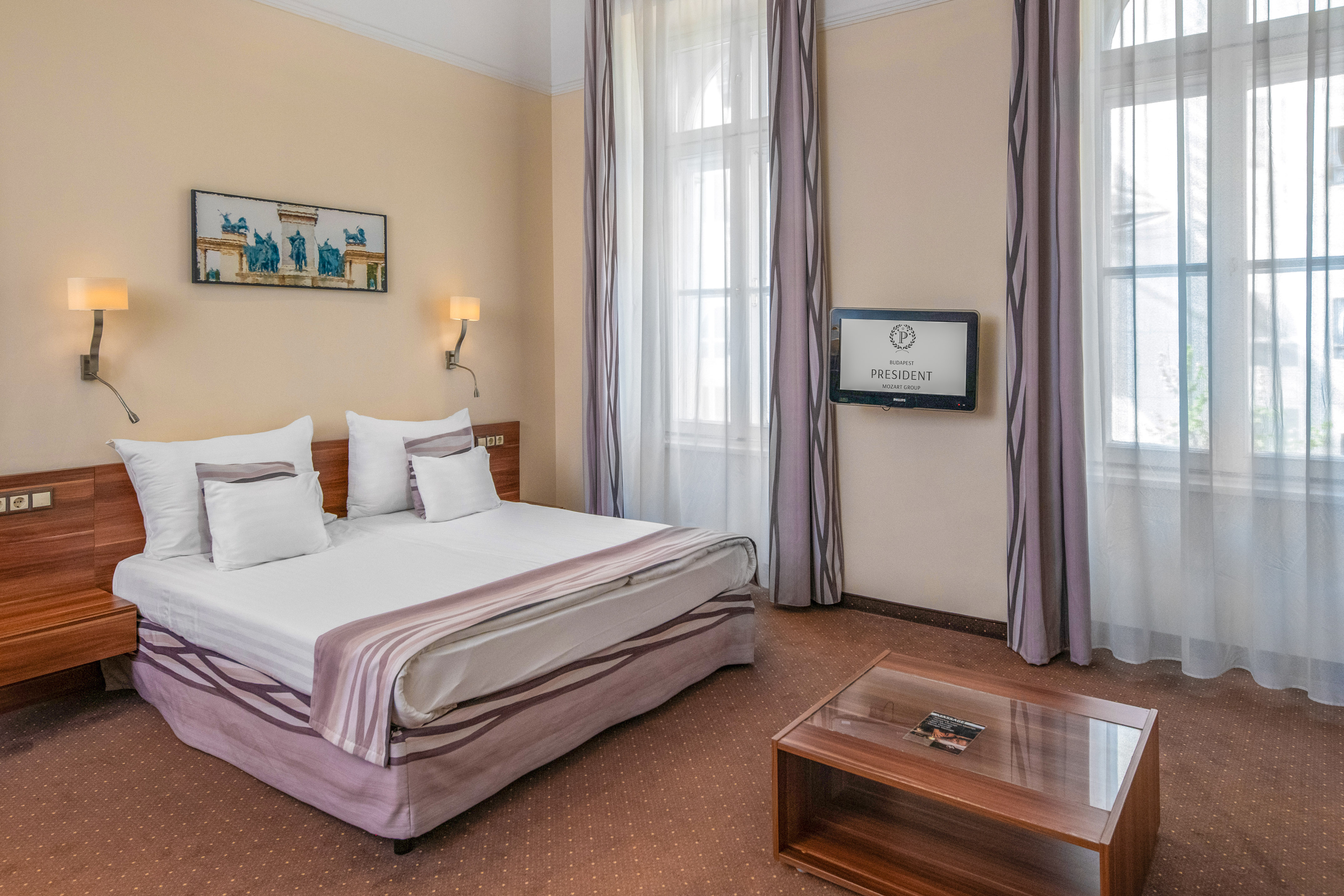 Hotel President Budapest bedroom with white bed, purple accents, windows, and TV.