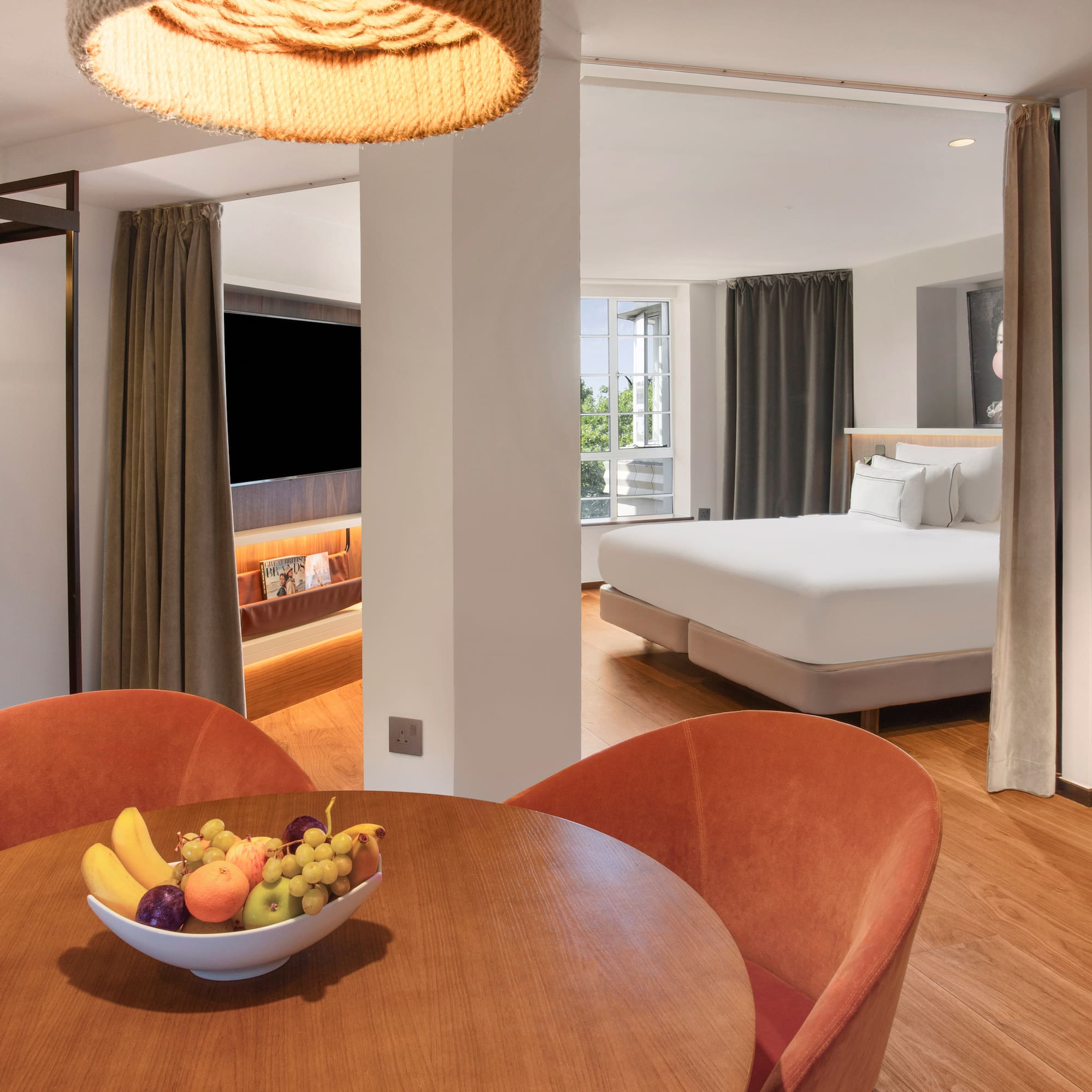 Modern hotel suite with dining area, fruit bowl, bathrobes, and comfortable bedroom.
