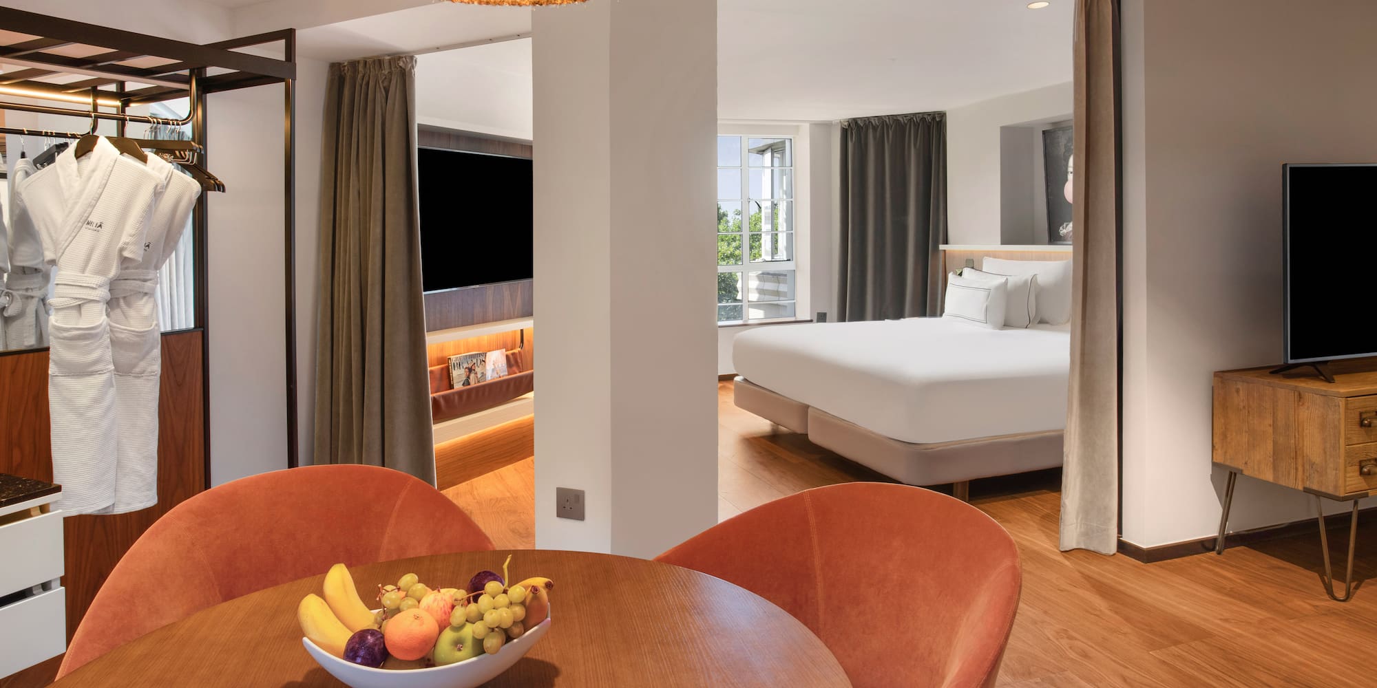 Modern hotel suite with dining area, fruit bowl, bathrobes, and comfortable bedroom.