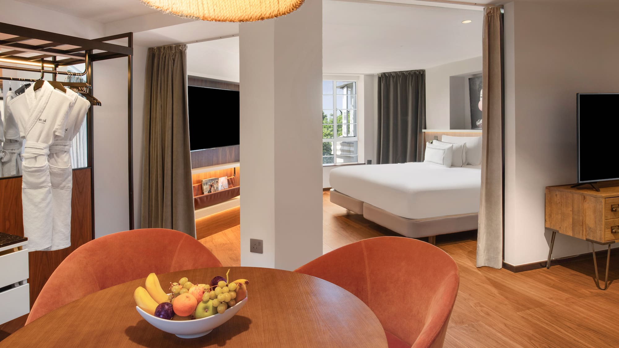 Modern hotel suite with dining area, fruit bowl, bathrobes, and comfortable bedroom.