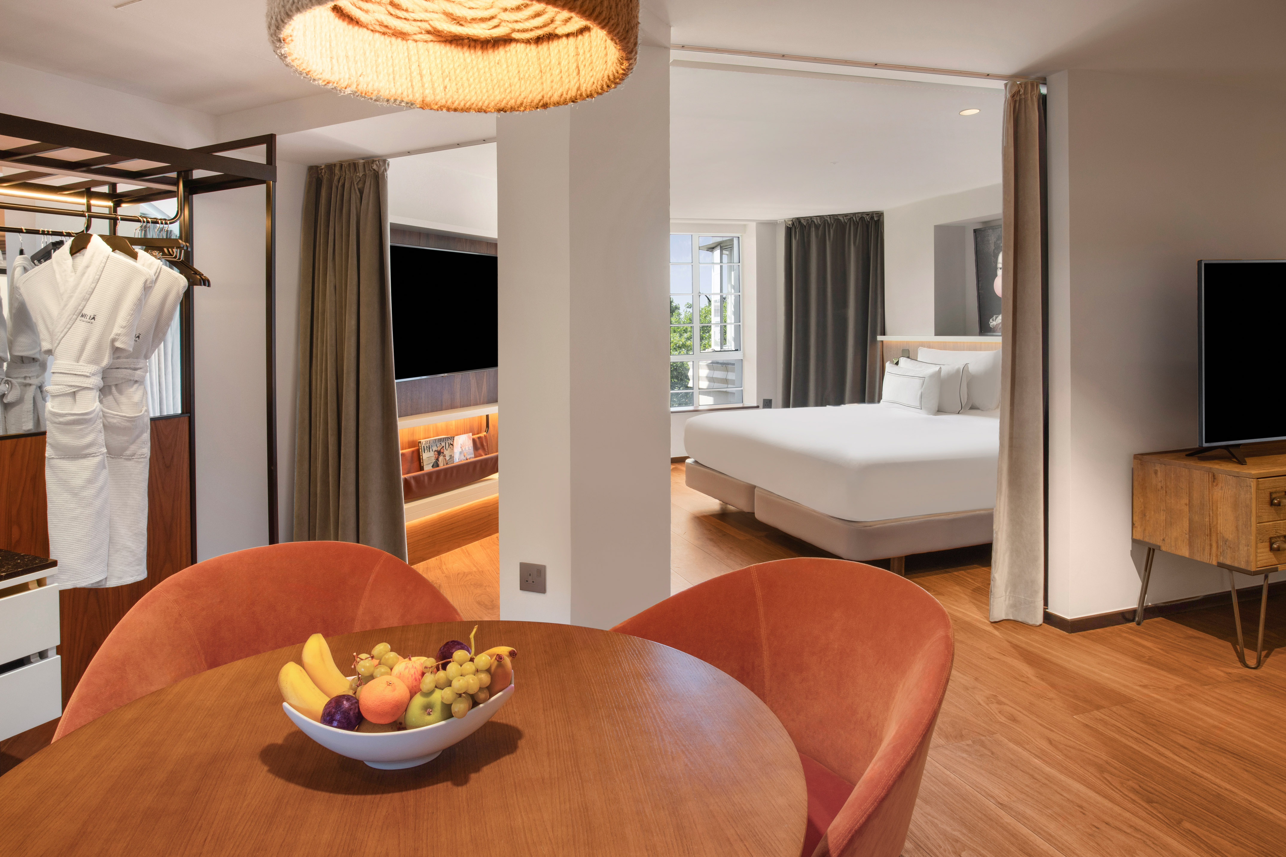 Modern hotel suite with dining area, fruit bowl, bathrobes, and comfortable bedroom.