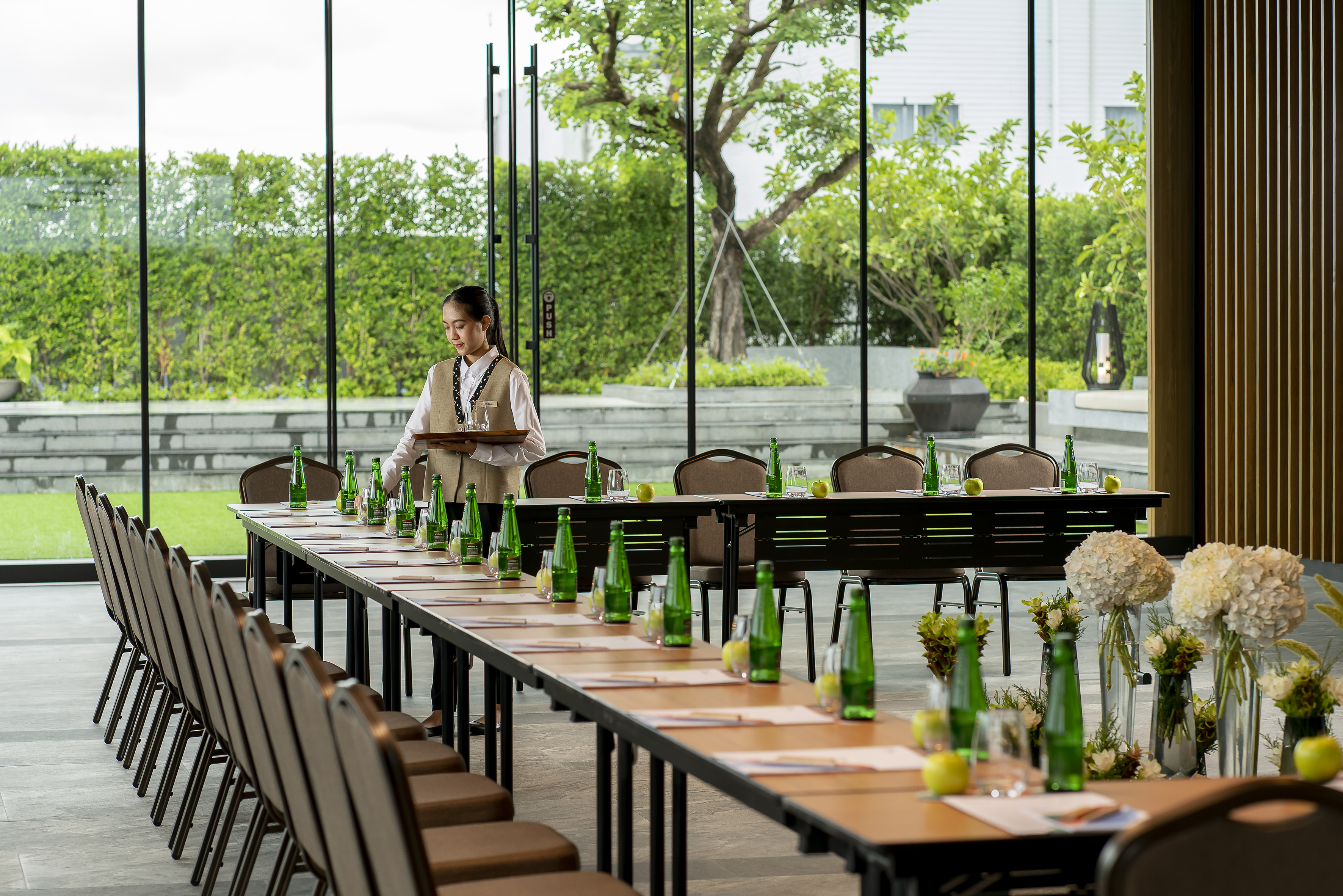 Server setting conference room with water bottles, apples, and green garden view.