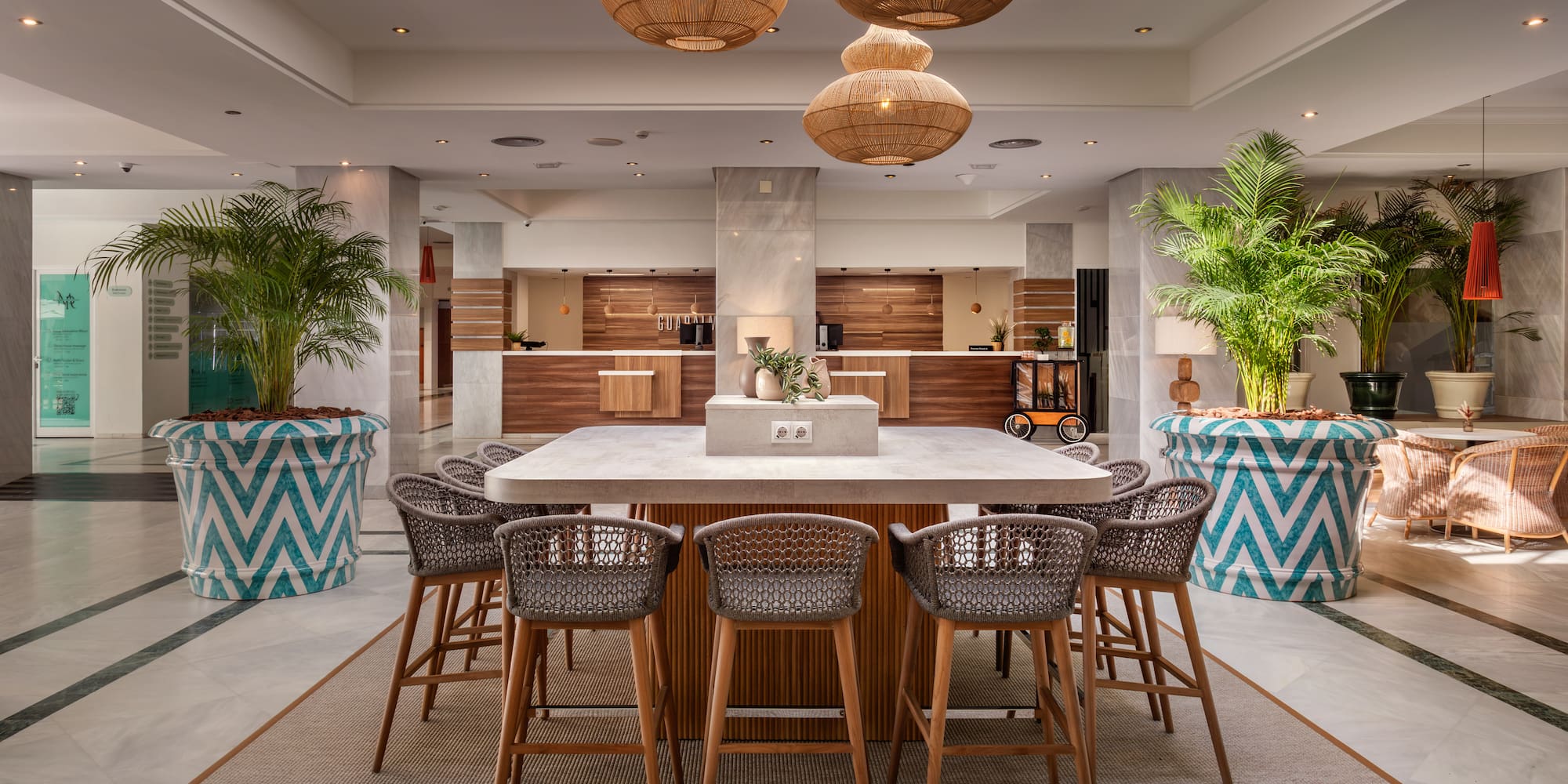 Modern hotel lobby with central communal table, reception, and rattan lights.