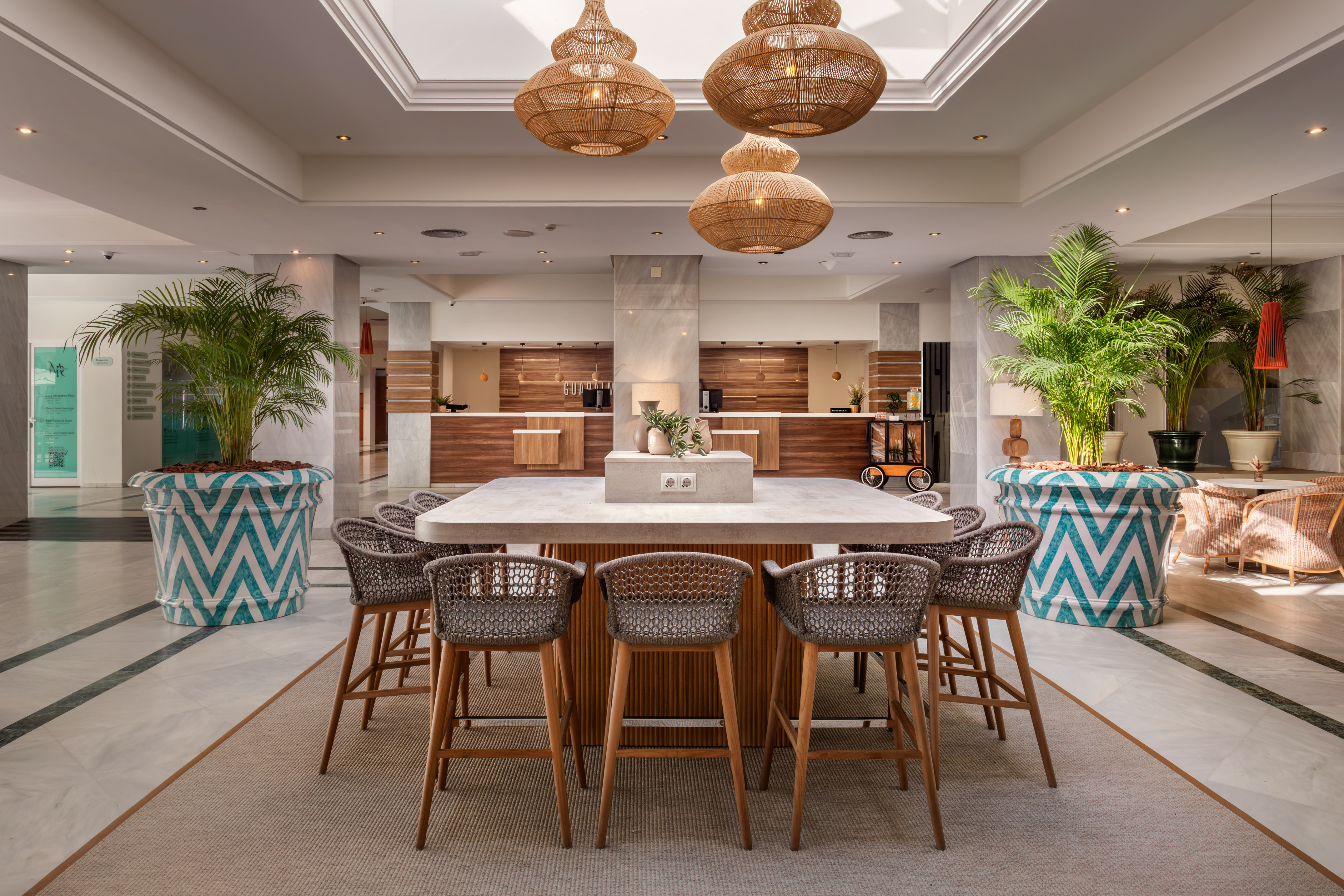 Modern hotel lobby with central communal table, reception, and rattan lights.