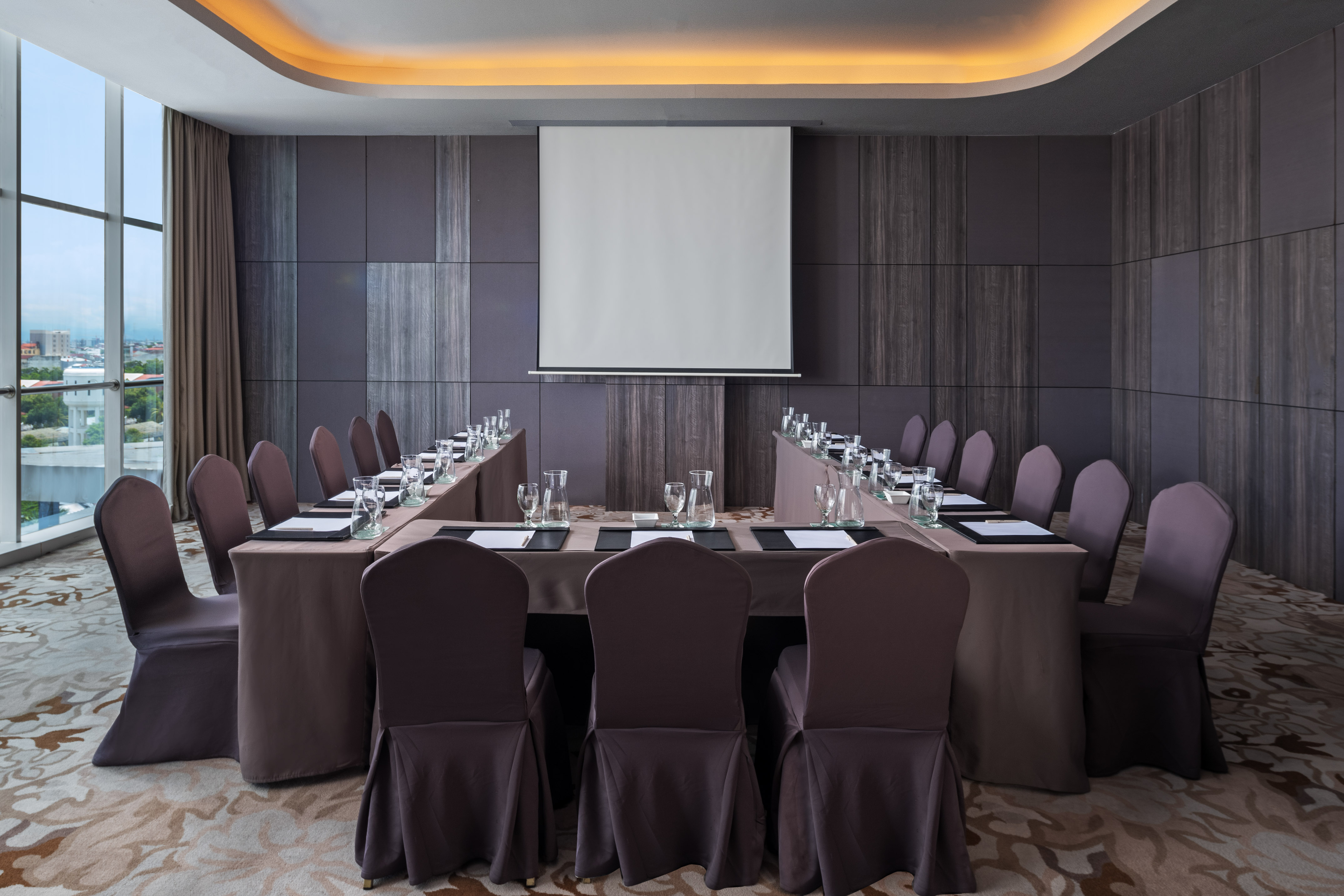 Modern meeting room with U-shaped table, projector screen, and city view.