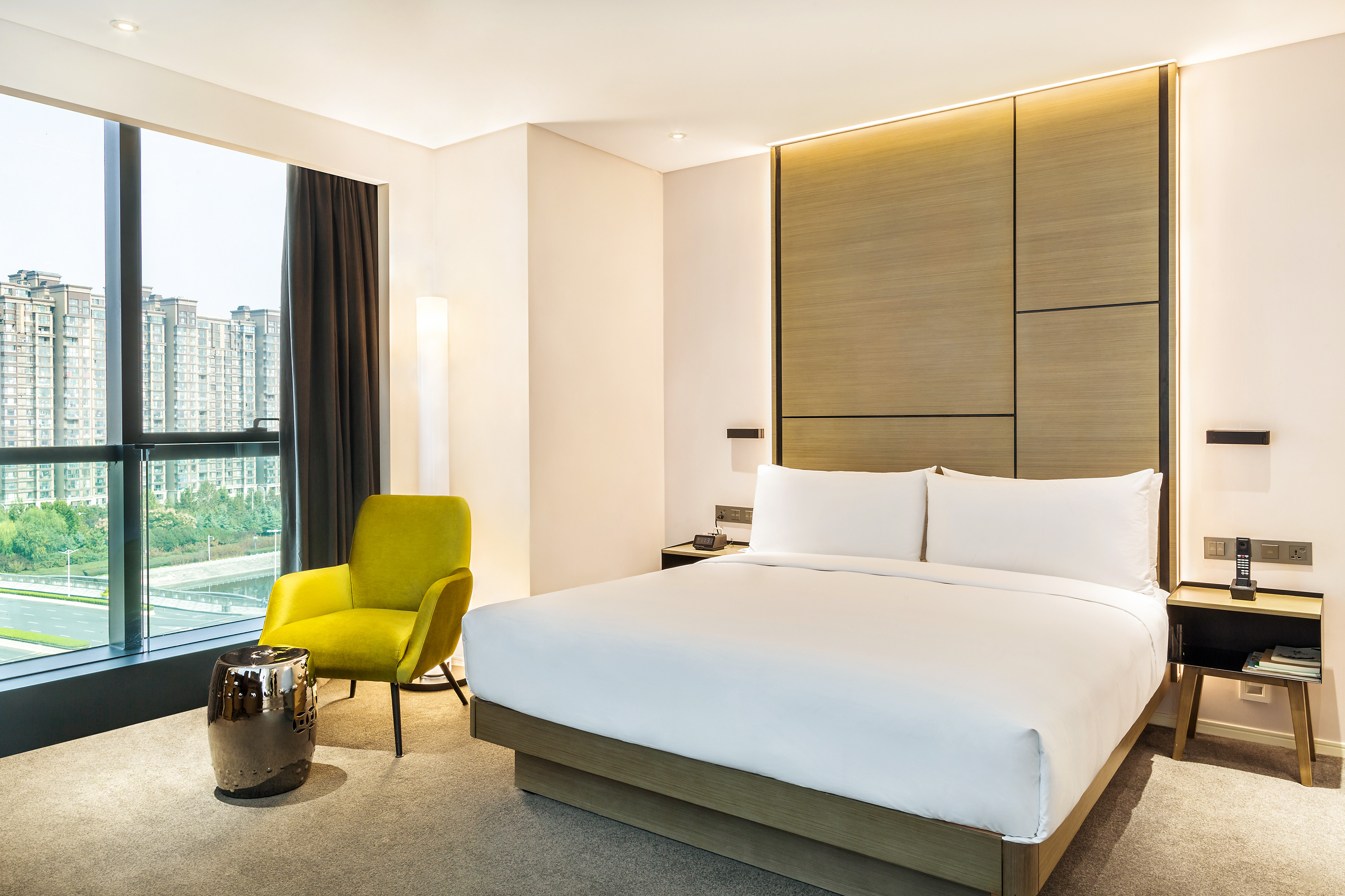 Modern hotel room with large bed, city view window, and chartreuse armchair.