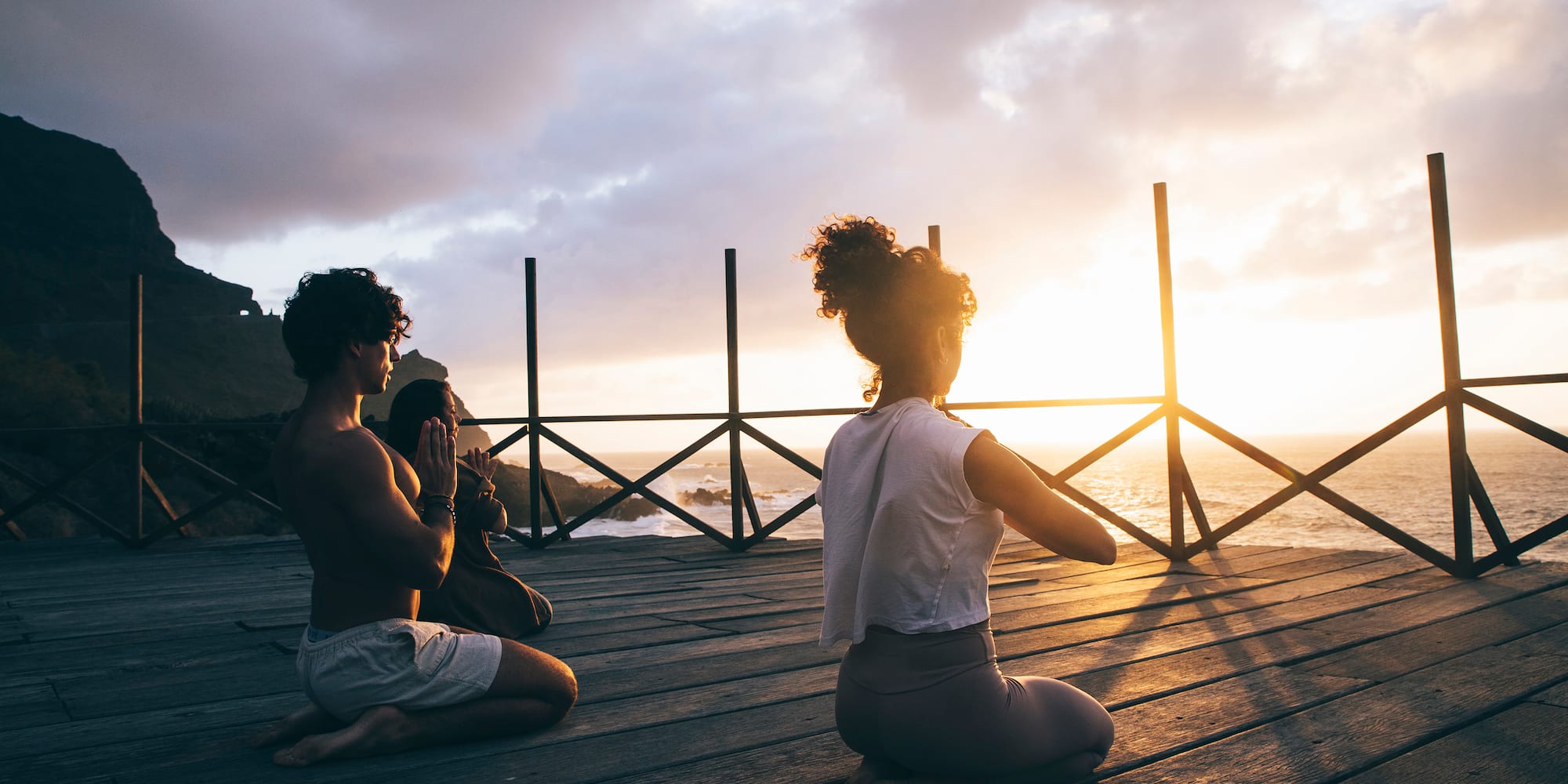 Meditating couple on wooden deck overlooking ocean at sunset.