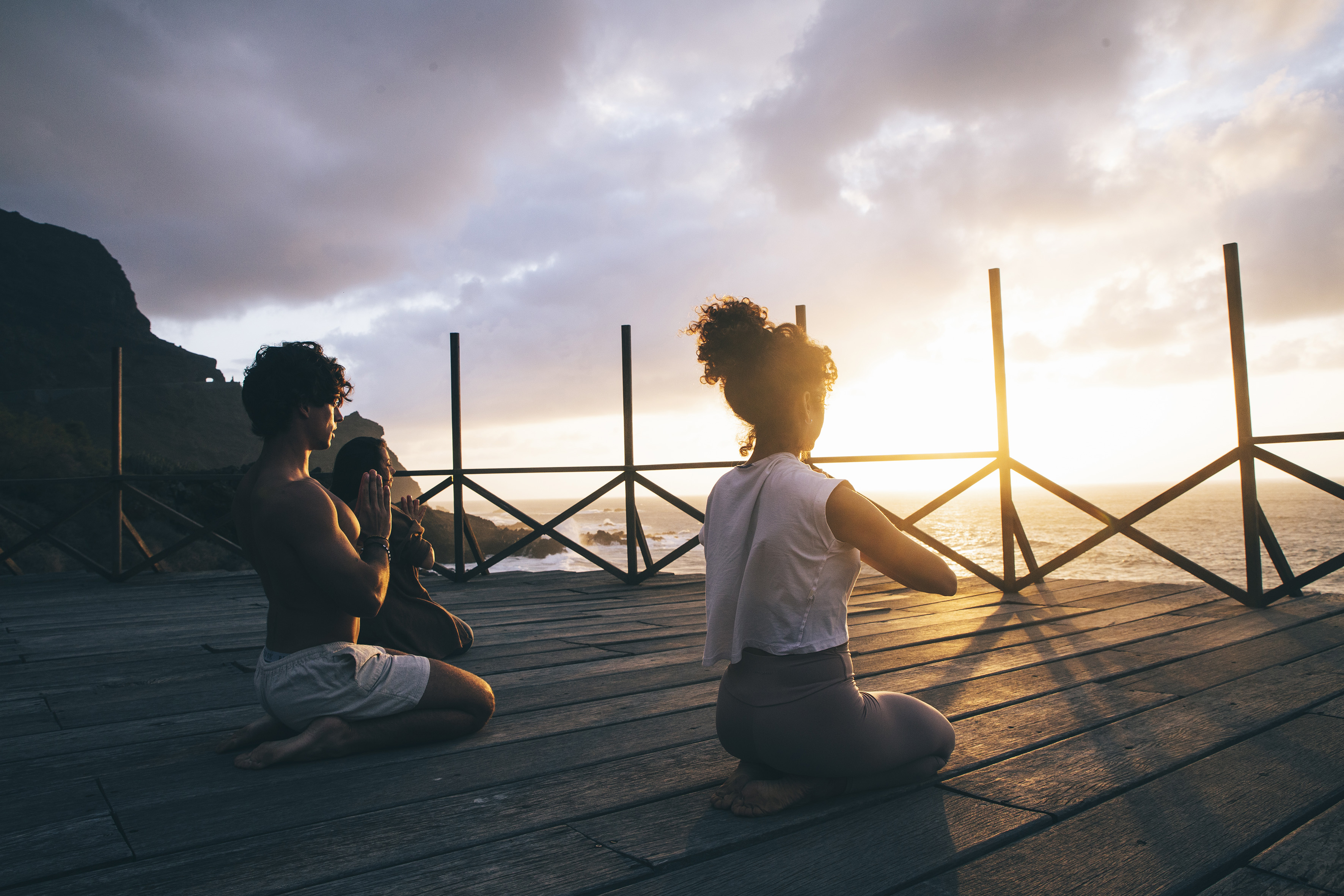 Meditating couple on wooden deck overlooking ocean at sunset.