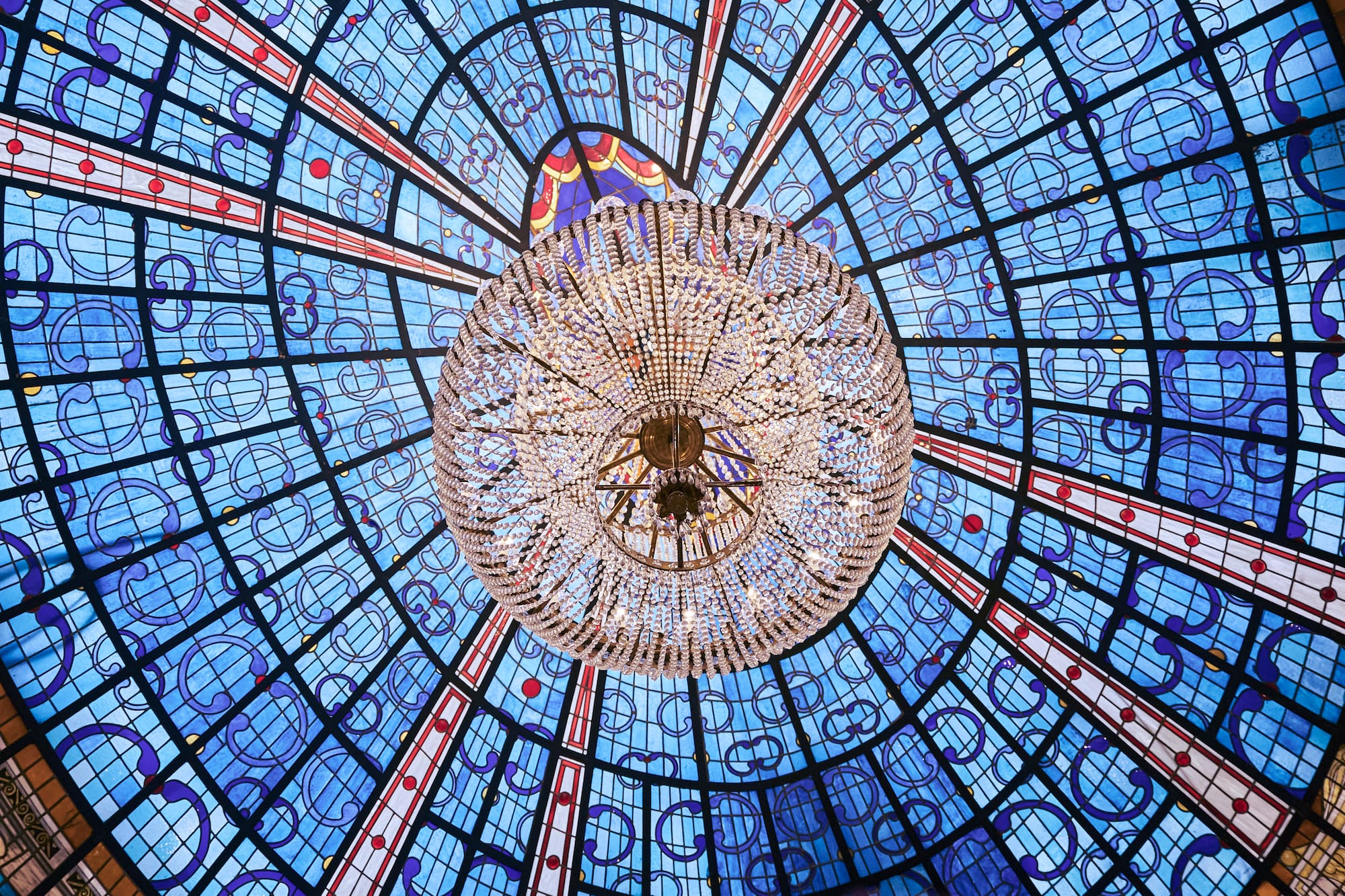 Ornate crystal chandelier under an intricate blue stained glass ceiling dome.
