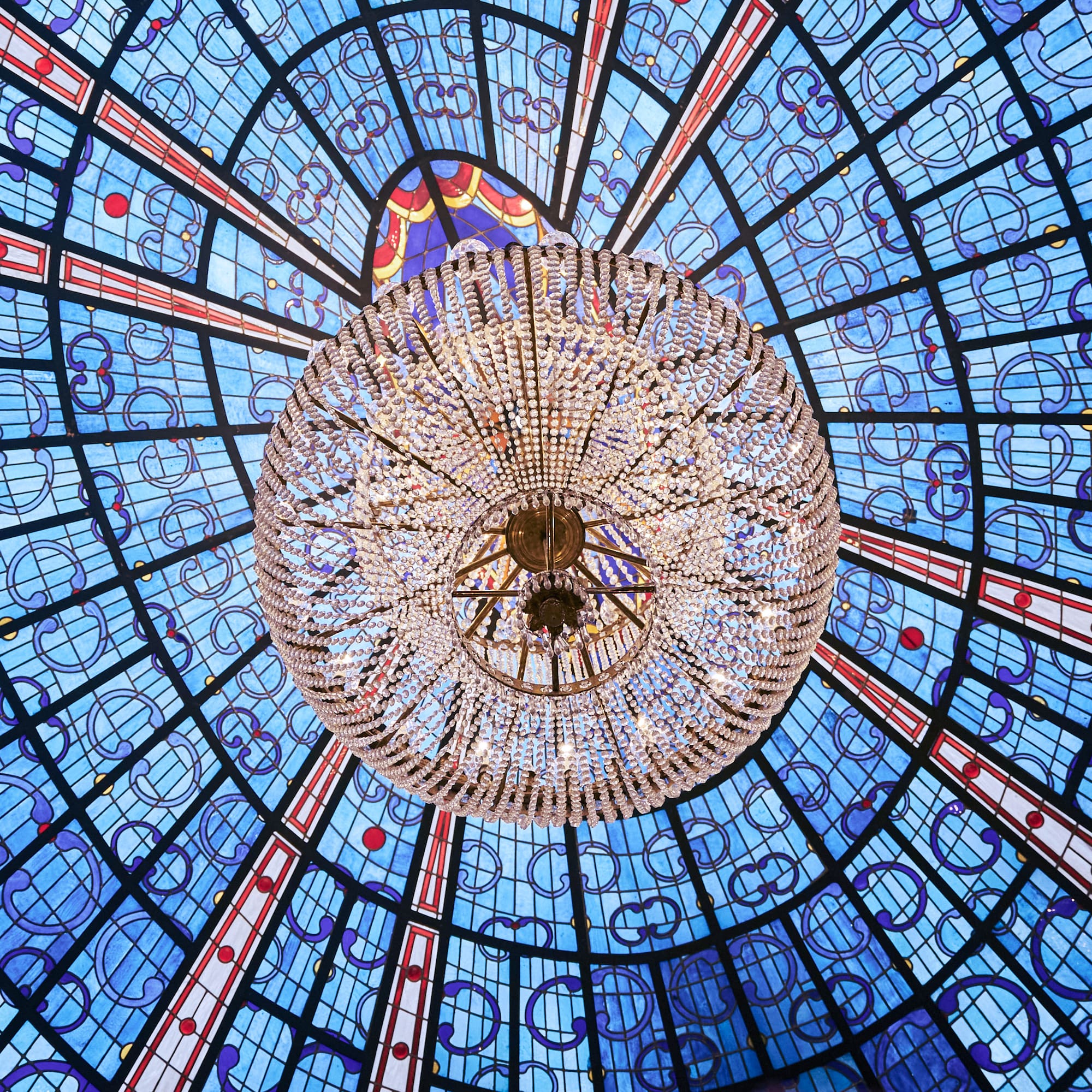 Ornate crystal chandelier under blue stained glass dome ceiling.